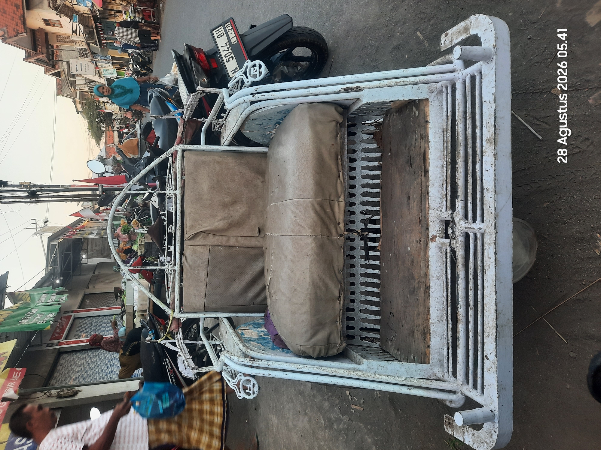 A three-wheeled motorized rickshaw, commonly known as a "becak motor," is parked on a paved road in front of shops. The becak motor is white and shows signs of wear, with peeling paint. It has a covered passenger seat and a wooden floor. A black motorcycle is parked behind the becak motor, displaying the license plate "N 5044 BH" and "11-28".
In the background, several people are visible. On the left, a man in a white and red striped shirt and a checkered sarong walks away from the camera carrying a blue plastic bag. Further down the street, a woman in a blue headscarf and a blue dress stands with a plastic bag. Other individuals are seen near shops and motorcycles. The environment appears to be a market or a busy street with shops lining both sides of the road. Flags and awnings are visible above the shops. The sky is clear, suggesting daytime. The scene is located in Pajarakan Kulon, Indonesia, as indicated by the timestamp "28 Agustus 2026 05.41".