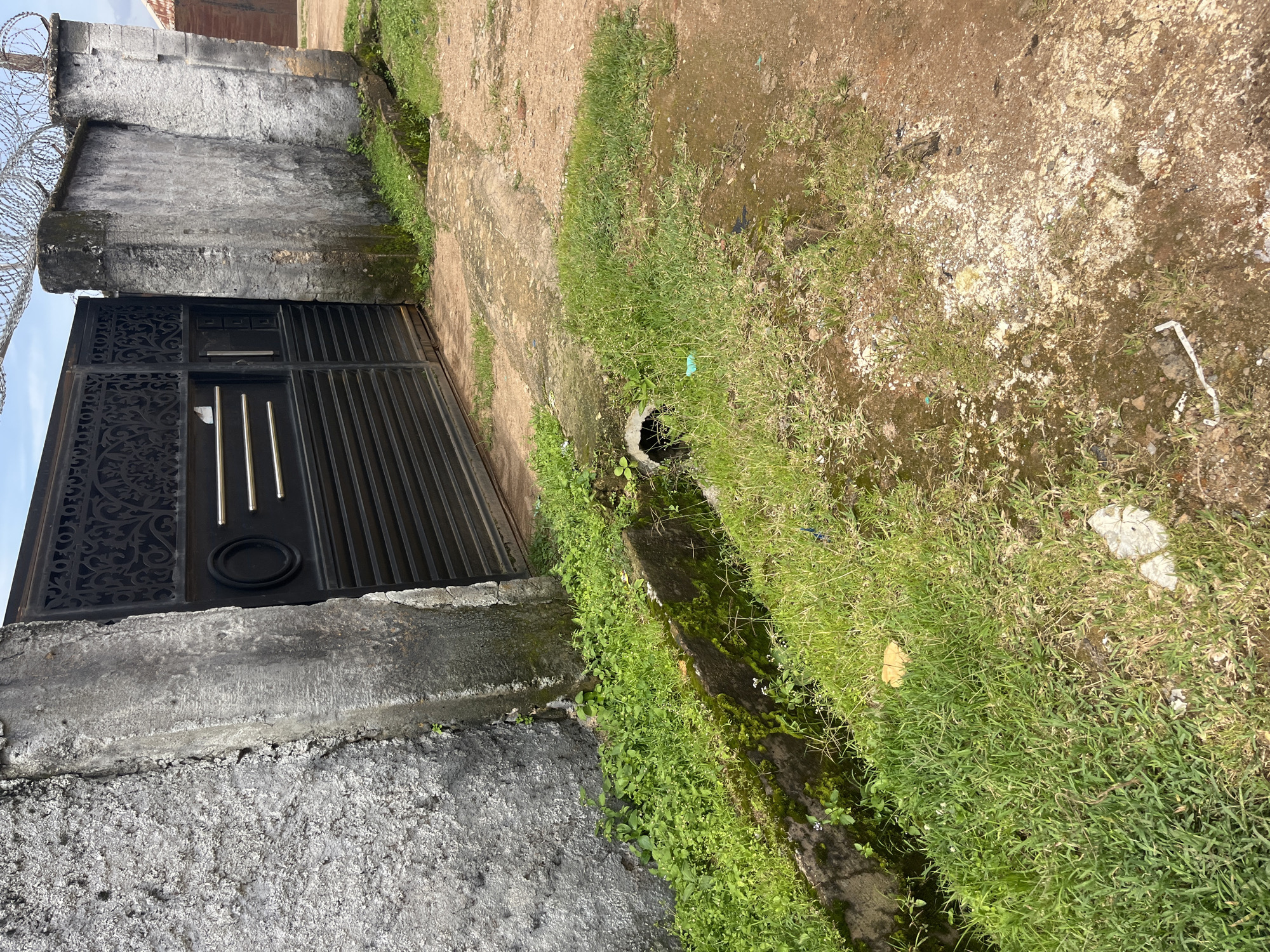 This image depicts an exterior view of a property with a large black metal gate and a concrete wall topped with barbed wire. A drainage ditch runs along the left side of the scene, overgrown with lush green grass and some moss. The ground is a mix of dirt and patches of grass, with some debris visible. The overall impression is one of a somewhat neglected urban environment. The scene is consistent with the typical streetscape found in many parts of Jos, Nigeria, characterized by a mix of concrete structures, unpaved surfaces, and natural vegetation.