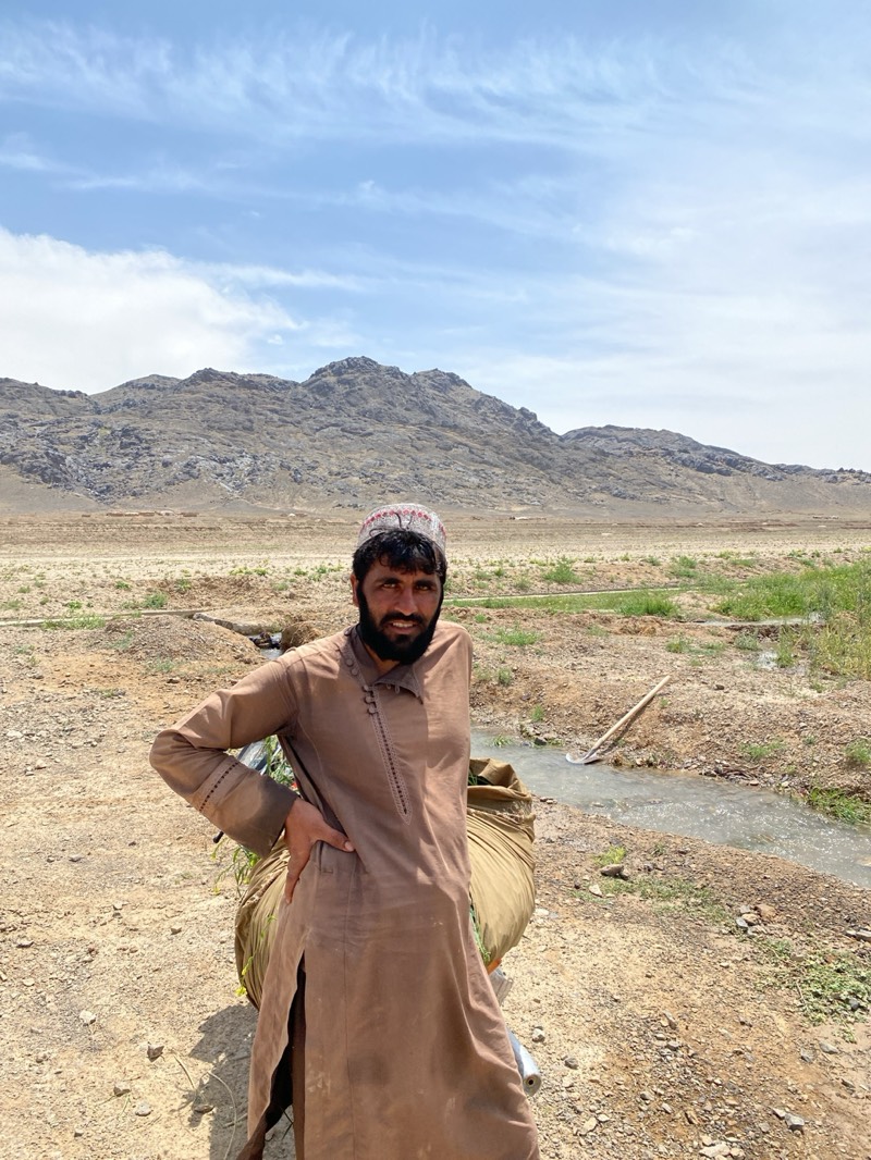 A medium shot captures a man in Ashoqah, Afghanistan, standing outdoors in a rural setting. He wears a brown, traditional garment with buttoned detailing at the neckline and a decorative cap. His left hand rests on his hip. A large sack is positioned behind him, appearing to contain plants.
The environment includes a dry, dusty foreground that transitions into a vegetated area with a small irrigation ditch carrying water. A hoe lies near the water. The backdrop reveals a landscape with arid mountains beneath a bright sky with scattered clouds.