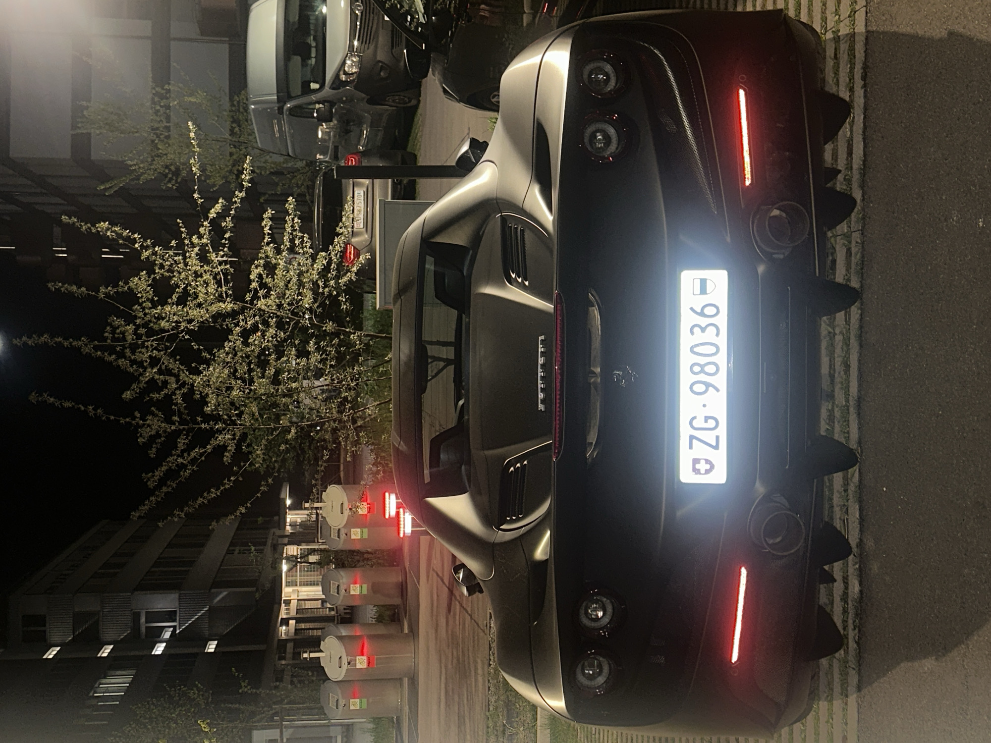The image captures a dark-colored Ferrari sports car, likely matte grey or black, viewed from its rear in an outdoor setting at night. The car is the primary subject, displaying distinctive round taillights, a long red light strip above the license plate, and quad exhaust pipes integrated into a prominent rear diffuser. A "Ferrari" badge is visible on the trunk lid. The Swiss license plate "ZG-98036" is brightly illuminated, indicating the canton of Zug.
In the background to the left, a vibrant tree or large bush with light-colored, possibly flowering or new spring leaves, stands out against the dark sky, illuminated by ambient light. Further back, multi-story buildings with some illuminated windows suggest an urban or suburban environment.
Other vehicles are partially visible in the background; to the upper right, a white van is parked, and below it, another car displays a Swiss license plate "LU-5675704" (Luzern). Along the left side of the frame, several light-colored, cylindrical structures resembling bollards or charging stations are visible, each emitting a red glow.
The scene depicts a static moment with no overt activity, suggesting the vehicles are parked. The artificial lighting from the car's lights, the bollards, and building windows highlights the nighttime setting. The ground appears to be asphalt with some grassy pavers.
