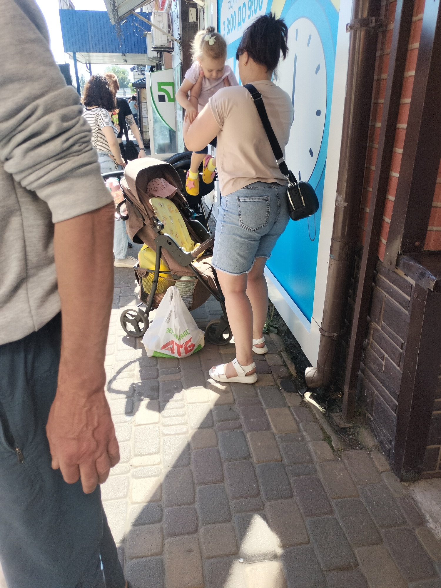 An adult woman wearing a light-colored t-shirt and denim shorts holds a young child. She stands next to a stroller with a baby blanket and a white plastic shopping bag on the ground beside it. A large blue clock face with white numbers and hands is visible on a wall behind them. A man's arm and shoulder are visible in the foreground, to the left of the frame, wearing a grey t-shirt and dark pants. The scene is set on a paved walkway under a covered structure, casting shadows from the sunlight. The image was captured on May 18, 2023, in Chernihiv, Ukraine.