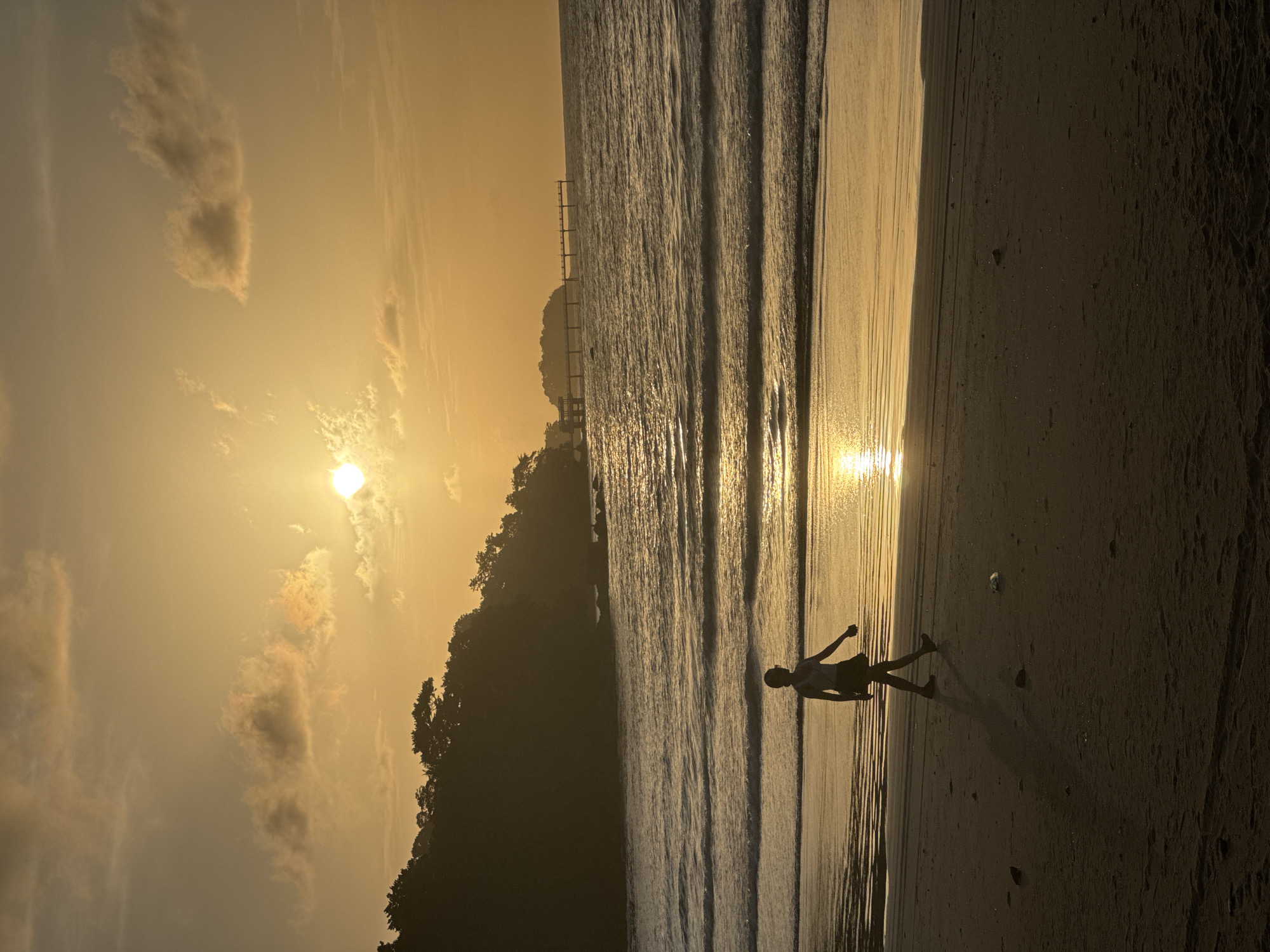 A person is walking on a beach during sunset. The sun is low in the sky, casting a warm glow over the scene. The waves are gently rolling onto the shore, and a pier extends out into the water in the distance. The person is silhouetted against the setting sun, and their shadow stretches out before them. The beach is sandy, and there are a few rocks scattered along the shoreline. The water is a deep blue, and the sky is a mix of orange, yellow, and pink. The overall mood of the image is peaceful and serene. The location is Patong Beach, Phuket, Thailand.