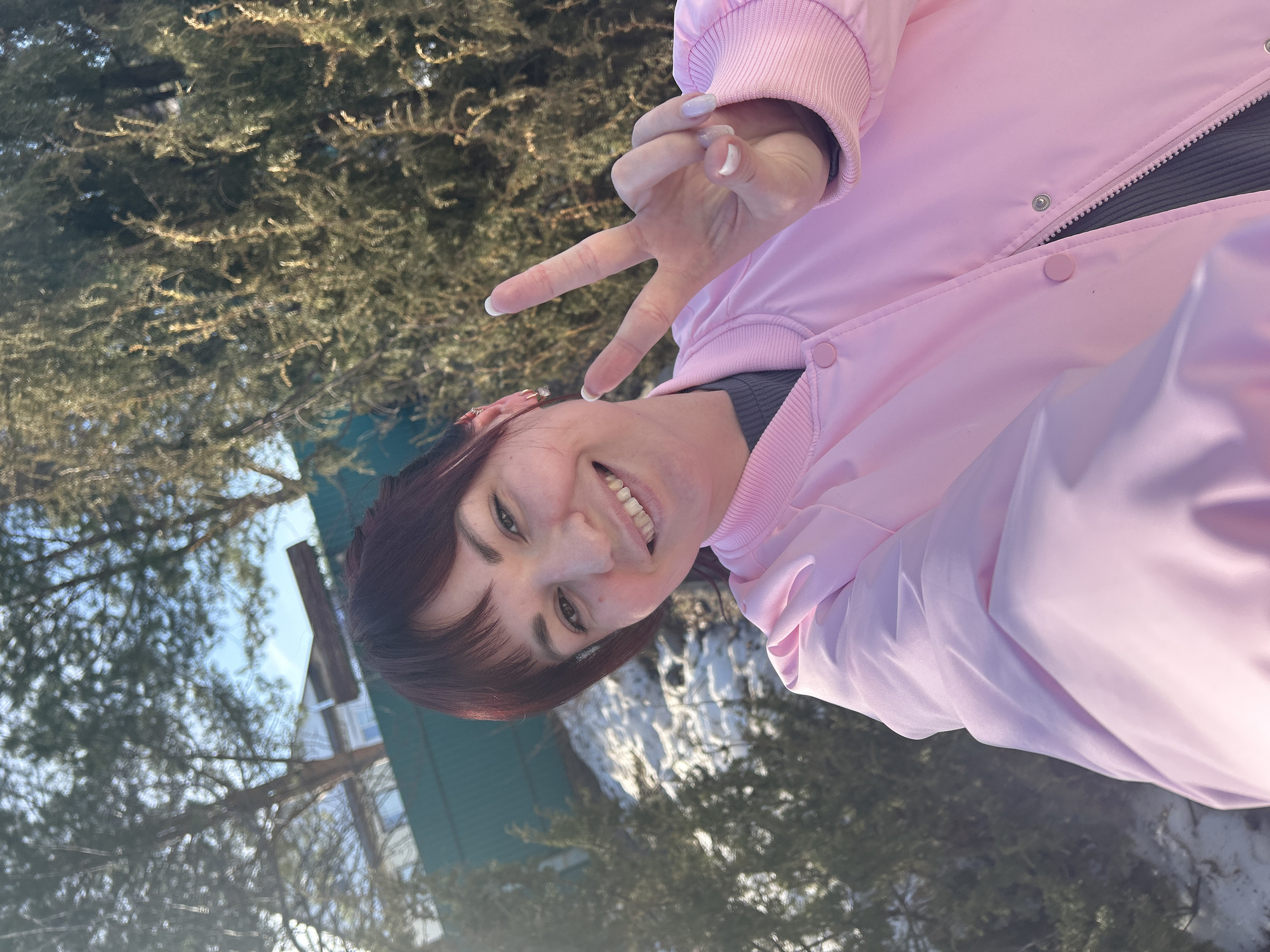 A woman in a pink jacket is outdoors, smiling and making a peace sign with her fingers. The background shows trees and a building with a green roof. Based on the prompt, the location is the city of Фоки, Russia. The presence of bare trees and what appears to be snow on the ground suggests it might be winter or early spring.