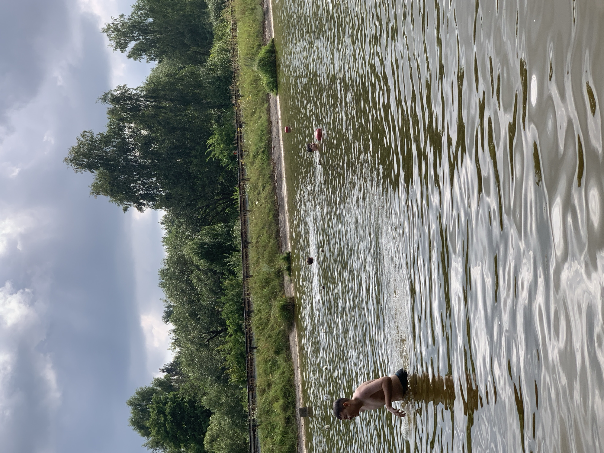 In the city of Грозаўскі сельскі Савет, Belarus, a boy is wading in murky water with ripples, appearing to be at the edge of a body of water. He is shirtless and wearing dark swim trunks. In the distance, another person is partially submerged in the water, and two red buoys are visible. The background shows a sandy shore with green grass and trees, and a dark, cloudy sky. The scene suggests a recreational activity on a somewhat overcast day.