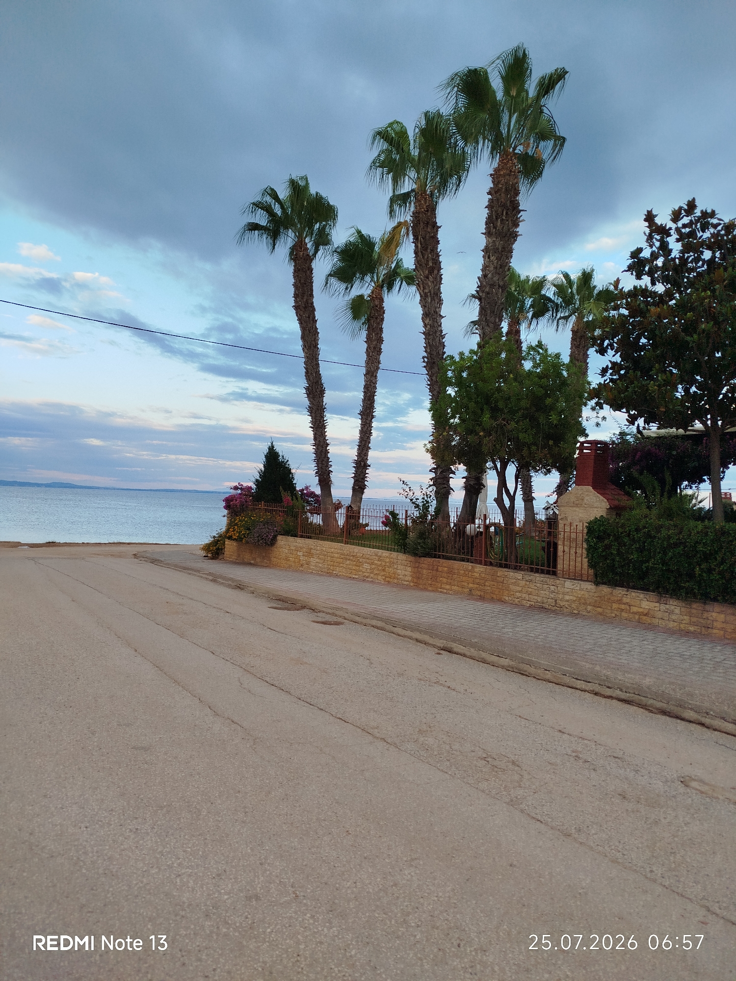A paved road runs diagonally across the foreground of the image, leading towards a body of water. To the right of the road, a low, light-colored stone wall borders a paved walkway. Beyond the wall, several tall palm trees stand silhouetted against a cloudy sky. There are also decorative plants with colorful flowers, lush green bushes, and a small brick chimney structure with a stone base. In the background, a calm body of water stretches to the horizon, with a faint landmass visible in the distance. The sky is a mix of blue and gray clouds, with hints of lighter clouds. The scene appears to be coastal, likely in a residential or resort area. The image is a photograph taken on July 25, 2026, at 6:57 AM.