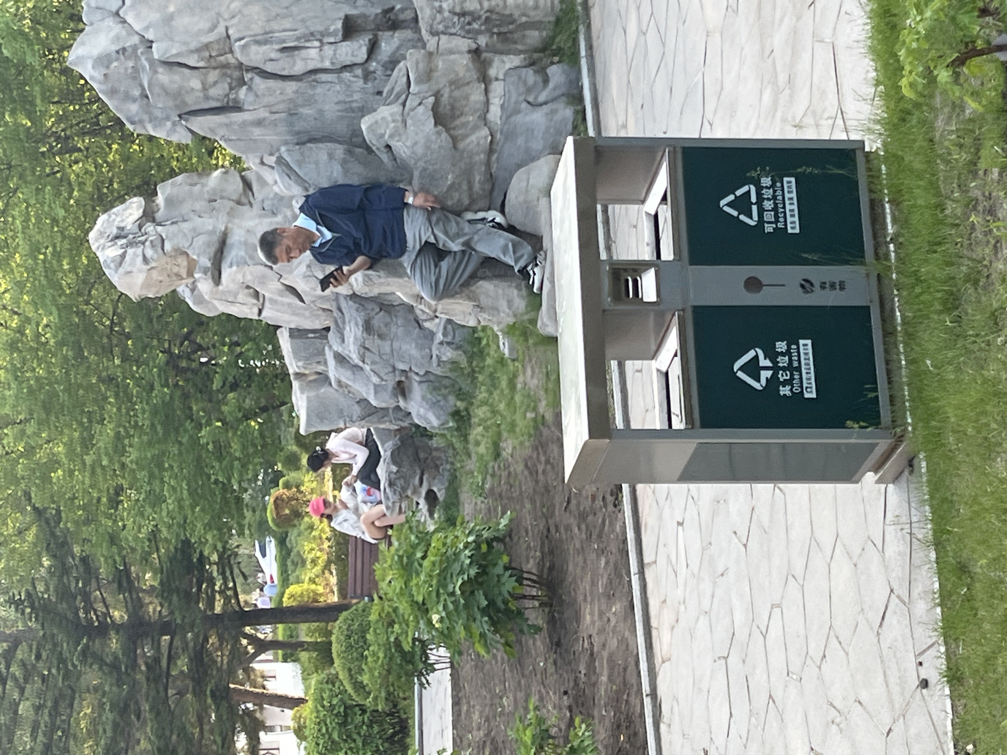 The image depicts a park scene in Changchun City, China. A man is reclined on a decorative rock structure, looking at a mobile phone. Nearby, two other individuals are seated on a bench. A dual recycling bin stands on a paved walkway. The surrounding environment includes trees, shrubs, and grass.