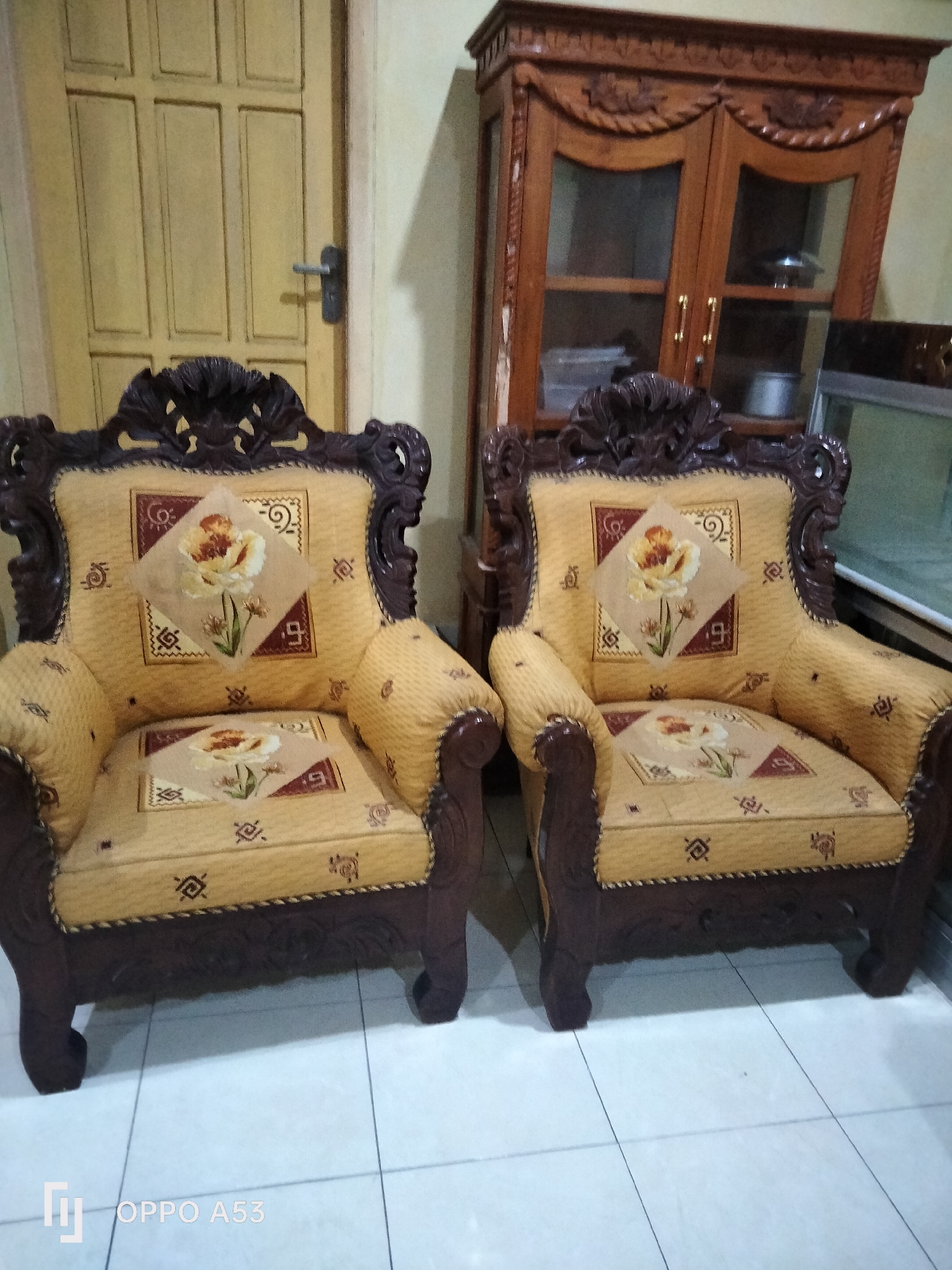 The image shows two ornate, upholstered armchairs with dark wooden frames. The upholstery is a light yellow fabric with a repeating pattern of small dark symbols and a central square motif containing a floral design. A small square throw pillow with a similar floral design is placed on each chair. Behind the chairs is a wooden cabinet with glass doors, and to the left is a yellow wooden door with a silver handle. The scene appears to be an interior setting, possibly a living room or reception area, with tiled flooring. The photo includes a watermark in the bottom left corner that reads "OPPO A53", indicating the device used for capture. There are no people or animals present in the image.