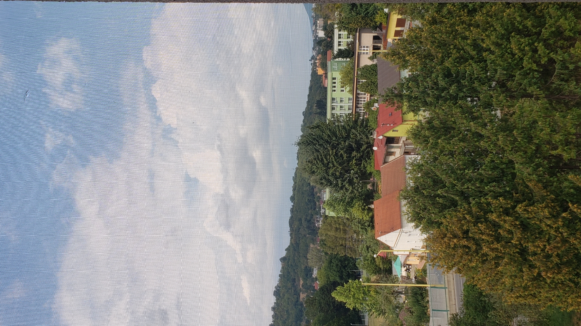 This is a daytime, medium-distance, outdoor, eye-level shot of a suburban neighborhood in Prešov, Slovakia, viewed through a window screen. The sky is partly cloudy with white clouds against a blue background. In the foreground, a dense row of evergreen trees with some yellowing foliage dominates the lower portion of the frame. Behind the trees, several residential houses with red and orange tiled roofs are visible, interspersed with yellow and white painted facades. Further in the background, a green multi-story building with many windows is prominent, along with other residential and commercial buildings and a wooded hill in the distance. A road with a yellow barrier is also visible in the mid-ground. No people are present in the scene.