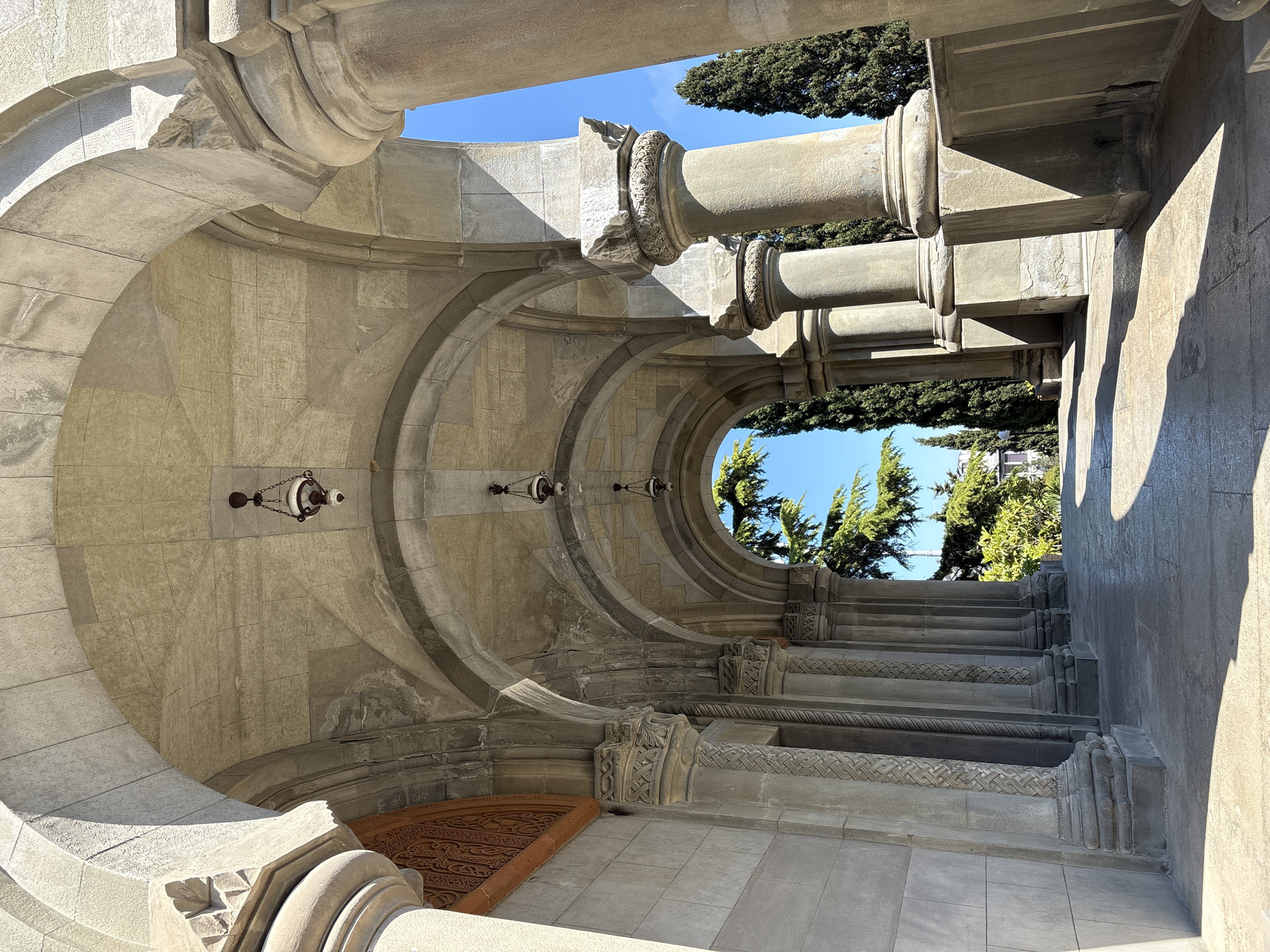 The image shows a stone archway and stairway in the city of Ялта, Ukraine. The archway is constructed of gray stone and features a large circular opening and smaller decorative arches with hanging lamps. Beyond the archway, there are trees and blue sky. The stairs are also made of gray stone and are decorated with carved details. Light and shadows play across the stone surfaces.