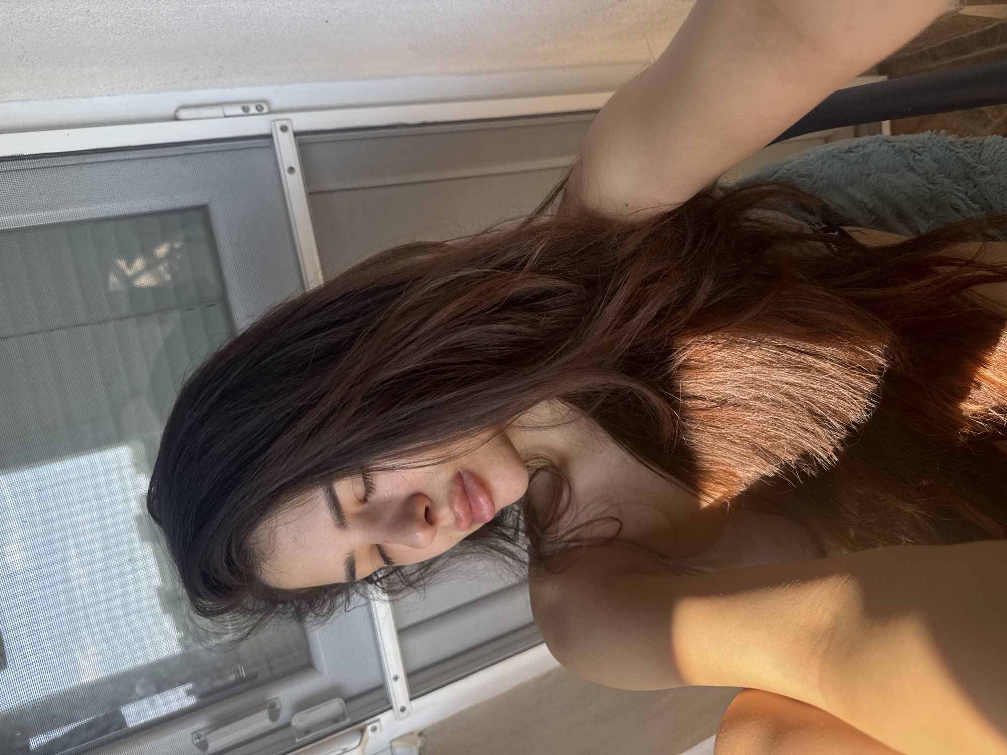 A close-up, high-angle indoor photograph depicts a young adult female in Plovdiv, Bulgaria, bathed in strong, direct sunlight. The subject has long, dark brown hair and fair skin, with her eyes closed and head tilted slightly to her left. Her lips appear full. She is bare-shouldered, with a subtle strap visible on her left shoulder, and parts of her left arm and upper left leg are illuminated by the sunlight in the lower-left quadrant. Her right arm extends out of the frame to the right, suggesting a selfie. The background features a white-framed window or glass door with a fine mesh screen, through which an indistinct outdoor environment with distant structures is visible. A textured, light-colored wall is present to the right of the window frame. A light blue/teal fuzzy object is partially visible in the lower-right corner, adjacent to a dark vertical furniture element.