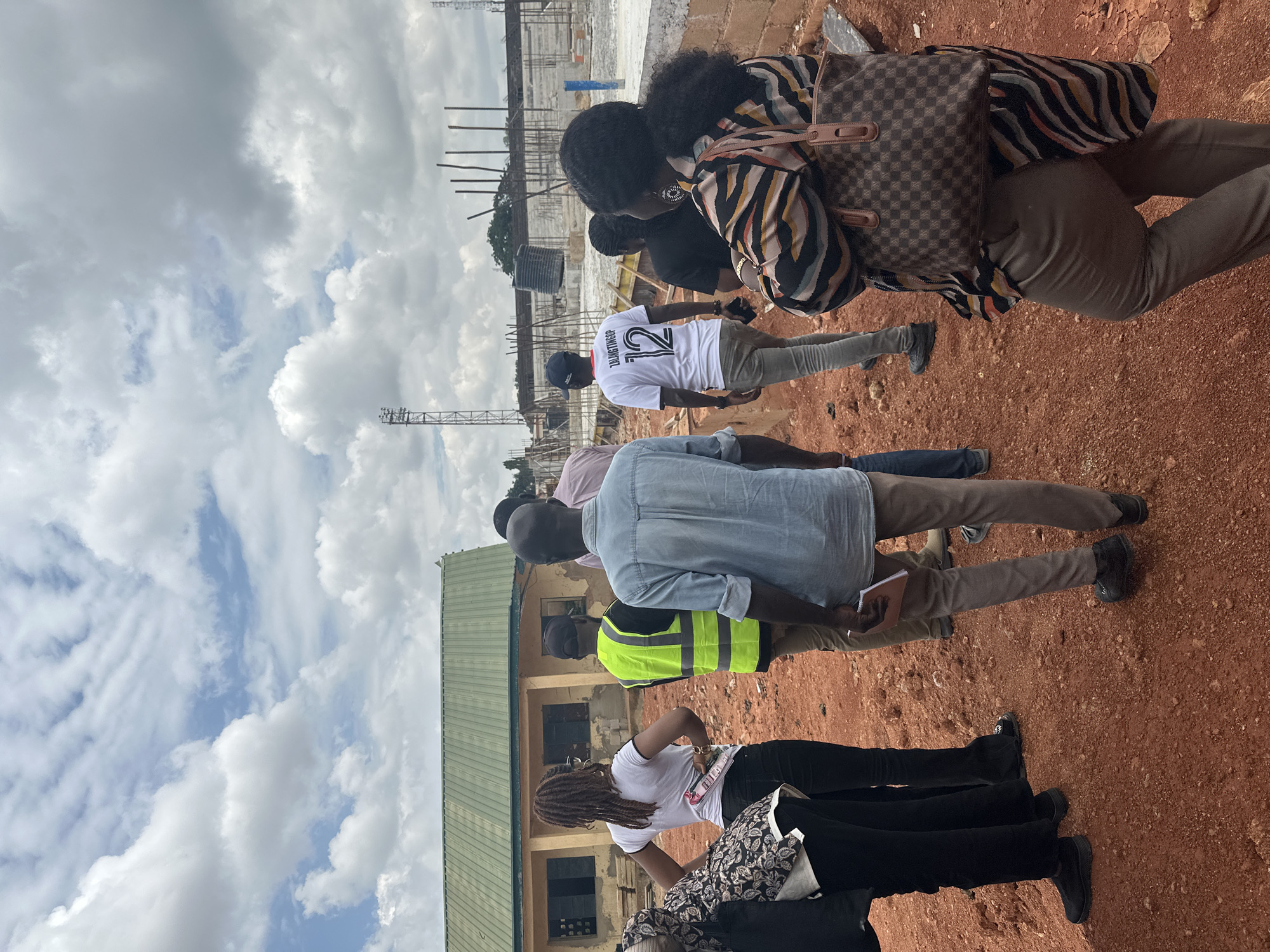 In Jos, Nigeria, on a construction site, a group of individuals stand on red earth. The sky above is filled with white, fluffy clouds against a blue backdrop. To the left, a building with a green roof and yellow walls is visible. In the background, unfinished construction structures rise, with rebar and scaffolding suggesting ongoing work. Several people are gathered, some facing away from the camera. One person wears a bright yellow vest, another a white t-shirt with "ZALANGTINGUP 12" printed on it, and a third wears a patterned top and carries a brown checkered handbag. The scene appears to be a site inspection or a meeting related to the construction project.
