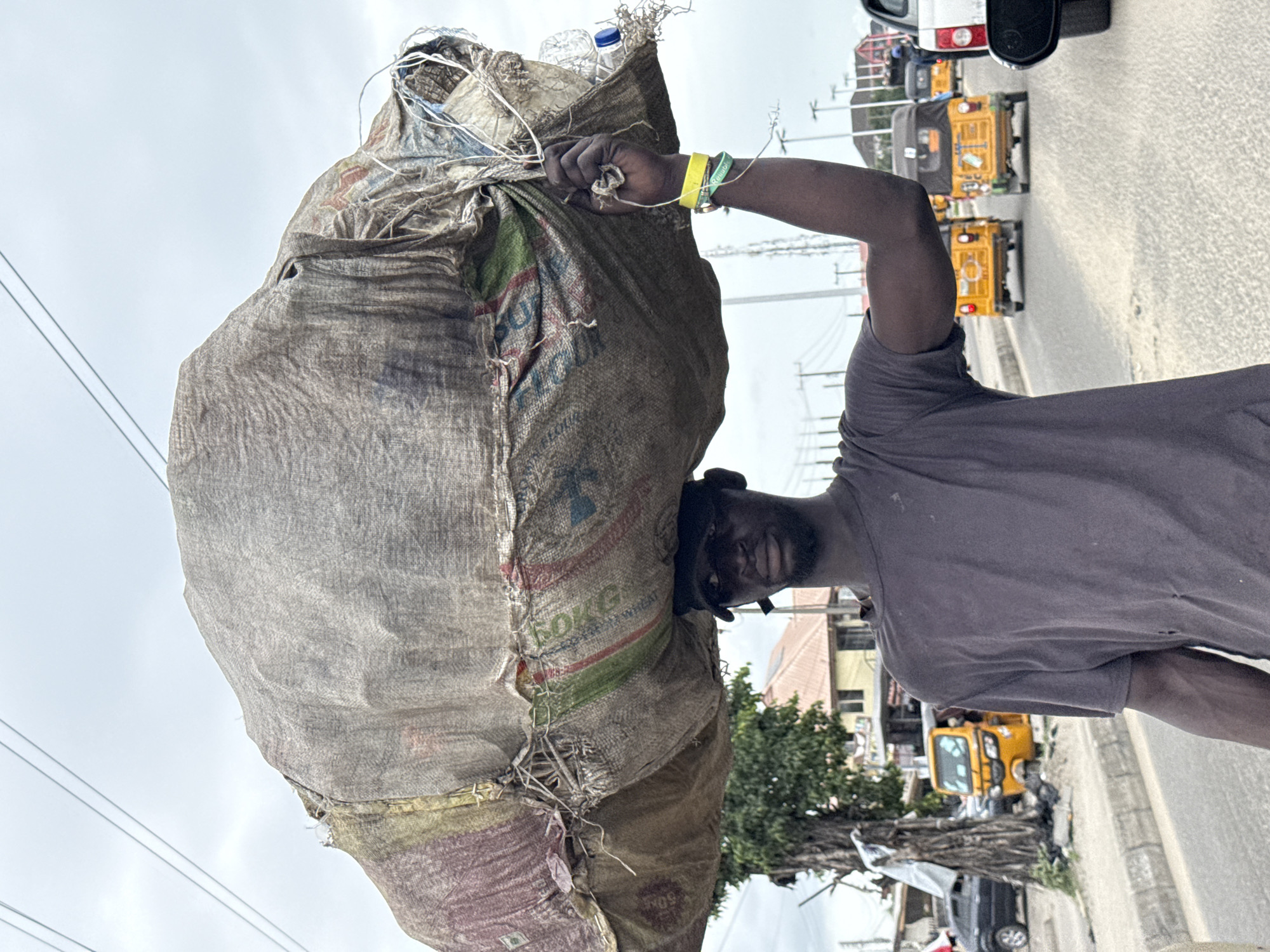 A man carries a large sack on his head, secured by his hand, on a street in Ado, Nigeria. He wears a faded gray t-shirt and has two wristbands on his right wrist, one yellow and one green. The sack is a rough, woven material, possibly jute, and appears to contain plastic bottles at the top. The side of the sack has a label with "50KG" and "WHEAT" visible. The man's face is partially obscured by the sack.
The background shows a street with several yellow and black three-wheeled vehicles ("keke napep") parked, as well as other cars. Utility poles with power lines are visible against a cloudy sky. A building and some trees can be seen in the mid-ground. The street itself appears to be paved and lined with a curb.