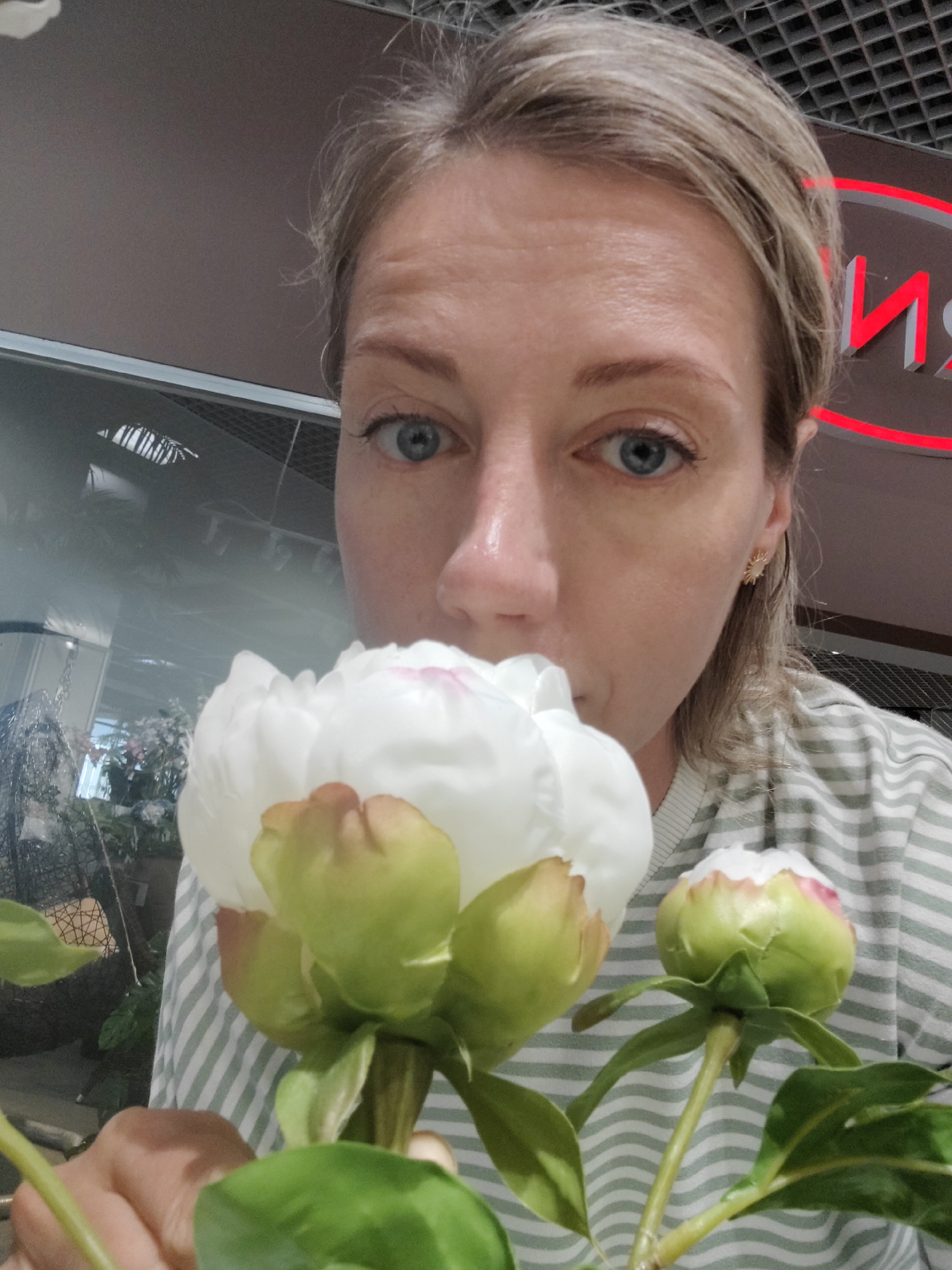 A woman with blonde hair is shown in a close-up, selfie-style shot, smelling white peonies. Her blue eyes are looking towards the camera, and her gaze is soft. She is wearing a green and white striped t-shirt and a small, gold, sun-shaped earring in her right ear. The background shows an indoor setting with a reflective surface, possibly a window or glass partition, through which greenery and decorative items can be seen. A red neon sign with Cyrillic lettering, which appears to read "ИМ", is visible in the upper right portion of the image. The lighting suggests it is daytime, and the overall mood is peaceful and focused on the sensory experience of the flowers. The location is identified as Saint Petersburg, Russia.