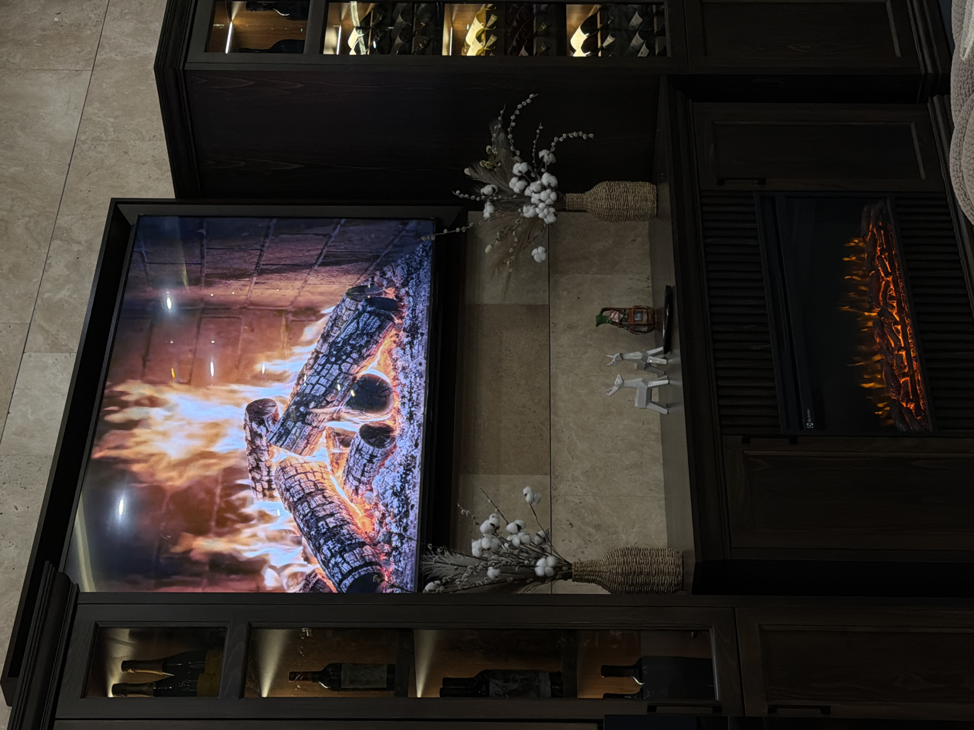 The image shows a television displaying a fireplace image, mounted within a dark wood entertainment center. Above the television are decorative elements, including a vase with cotton stems. To the right is a faux fireplace set within the center. Beneath the television are glass-fronted cabinets housing wine bottles. The scene is indoors. No location in Tsakhkadzor, Armenia is specified or implied by the image.