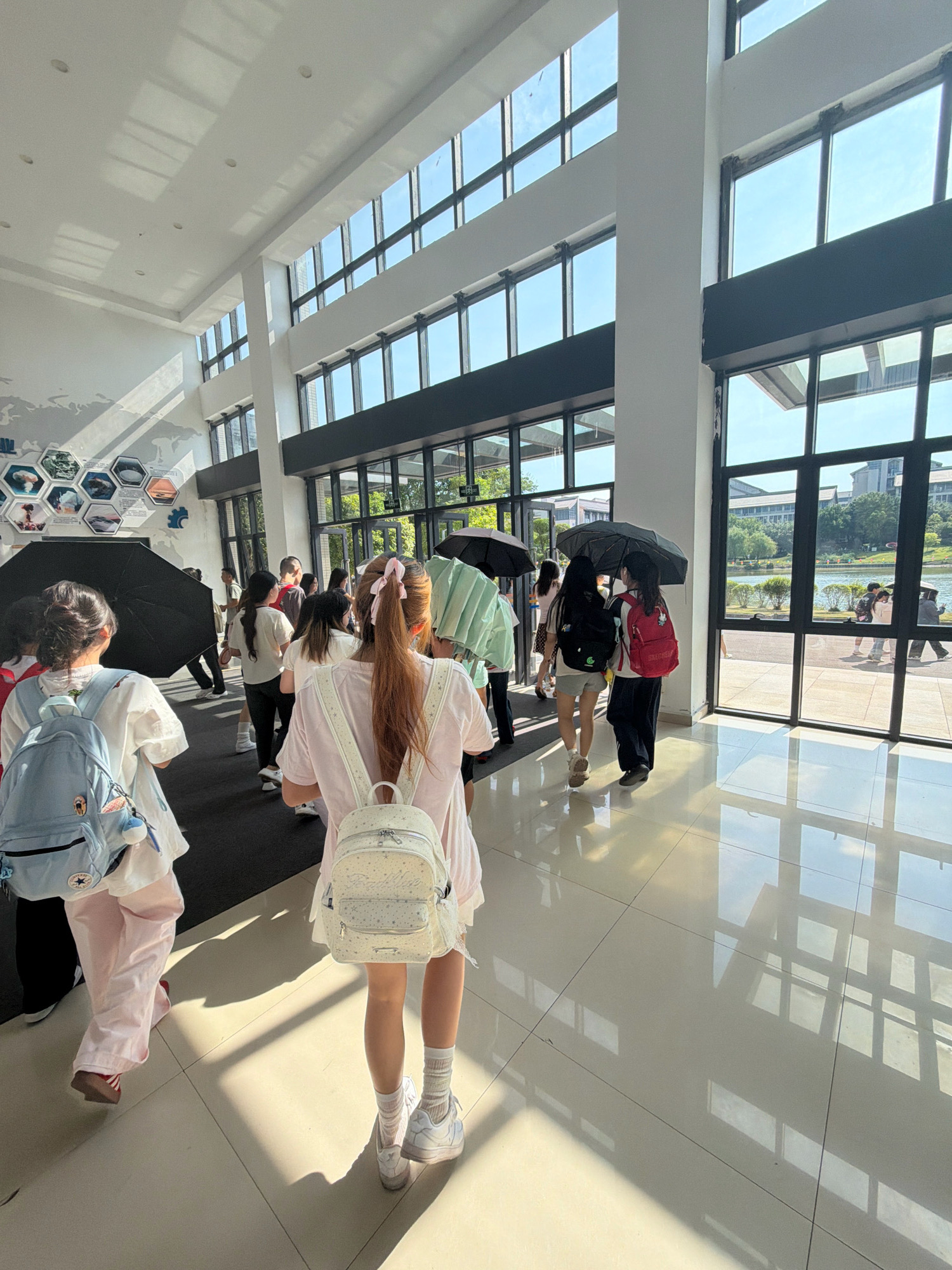 In a sunlit, modern interior lobby with a high ceiling and large windows, several individuals, primarily young women, are walking. The floor is tiled and highly reflective, showing patterns of sunlight and shadows from the windows. Several people are carrying umbrellas, suggesting it might be sunny or there's a possibility of rain. The environment appears to be an academic or public building, with displays on the wall featuring hexagonal frames containing images. Through the windows, a body of water and trees can be seen, indicating an outdoor environment beyond the building. The image captures a moment of movement and activity within this spacious architectural setting. The presence of backpacks suggests that the individuals may be students or attendees of an event. The general atmosphere is bright and open, illuminated by natural light. The location is identified as Jiangjin, China.