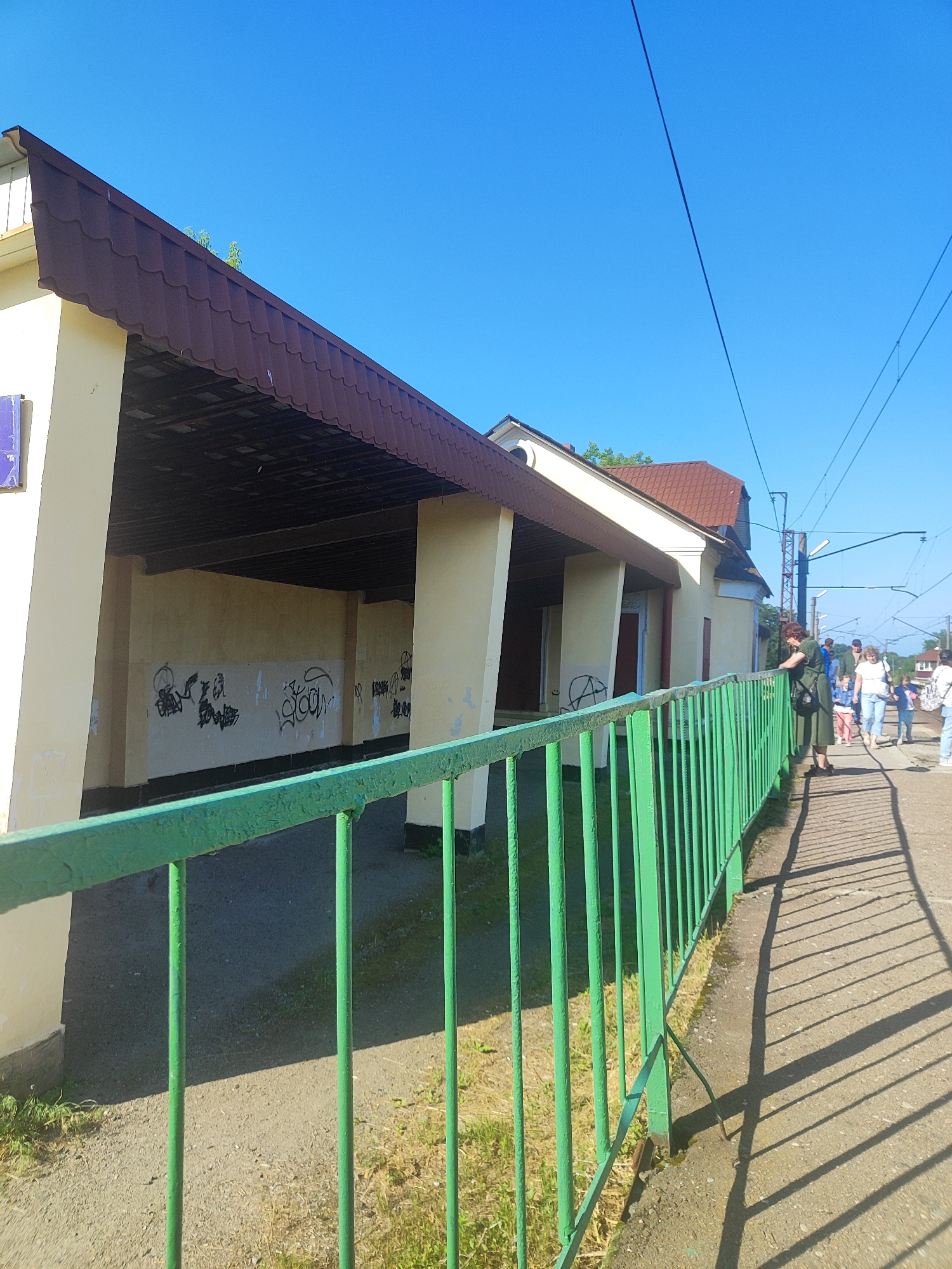 This is a daytime, eye-level, outdoor, full shot image of a train station platform with a building to the left and a walkway to the right. The building, painted a pale yellow, has a brown, tiled roof and appears to be a covered waiting area. The interior walls of the waiting area are covered in black graffiti. A bright green metal fence runs along the foreground, separating the waiting area from the walkway. Several people, including men, women, and children, are walking on the concrete walkway towards the right of the frame. The walkway is cast with long shadows from the fence and overhead power lines which are visible against a clear blue sky. The ground on the platform is a mix of concrete and dirt.