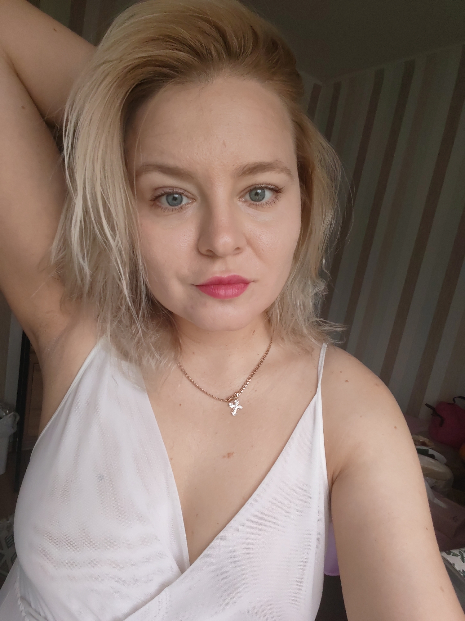 This is a selfie of a young Caucasian woman with blonde hair and blue eyes, wearing a white dress and a silver necklace with a cross pendant. She is indoors, with a striped wall in the background. The image does not contain any information that would allow for the determination of her location in the city of Izhevsk, Russia.