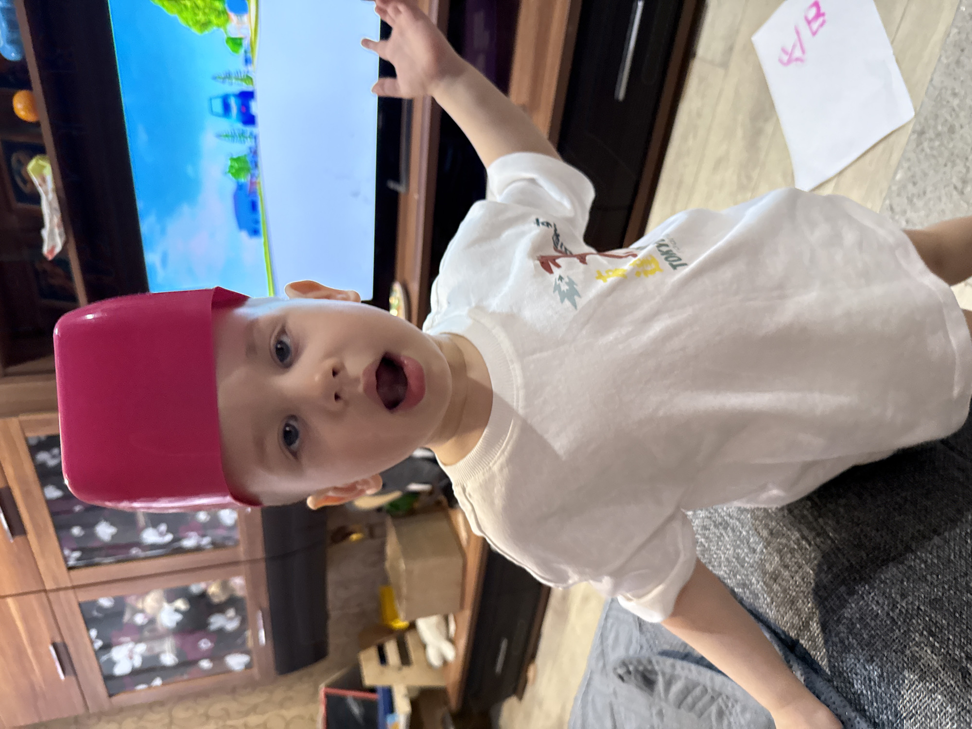 A child with a pink container on their head is looking at the camera with their mouth open and one arm raised. The child is wearing a white t-shirt. Behind the child is a television showing a cartoon. In the background is furniture and a paper with writing on it. The location is indoors, possibly a residence. Location information (Rusanovo, Russia) is not discernible from the image.