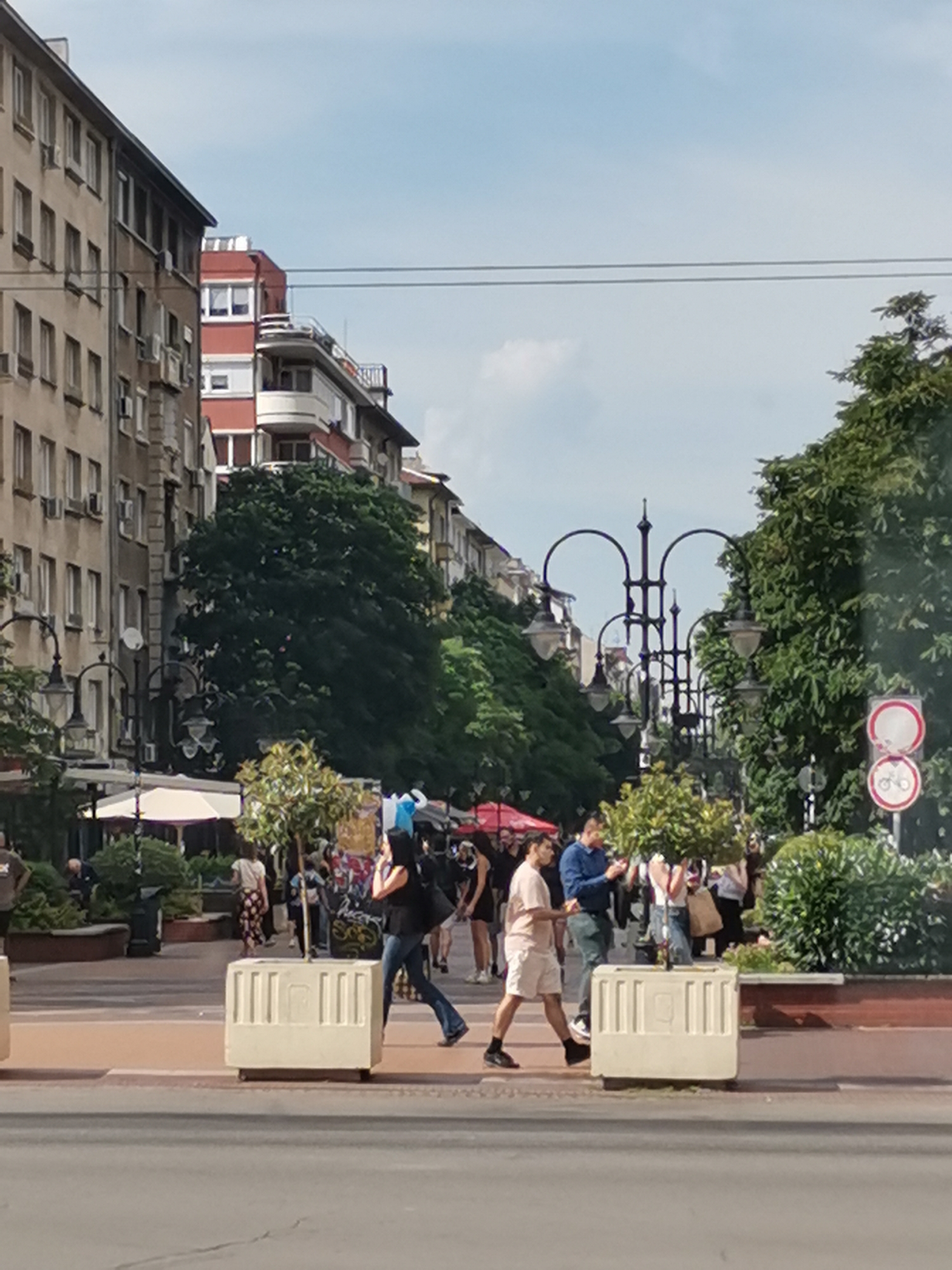 The image shows a street scene in Sofia, Bulgaria. In the foreground, there are large white concrete planters placed on the sidewalk. The planters have vertical grooves. Beyond the planters, several people are walking. Some are casually dressed, while others appear to be shopping or browsing. There are trees lining the street and buildings in the background. The architecture of the buildings suggests a European city. One of the buildings has a red and white facade. There are also streetlights with multiple lamps. On the right side of the image, there are traffic signs, including one that appears to indicate a bicycle crossing or a no-cycling zone. The sky is partly cloudy. The overall impression is a busy urban street with pedestrian activity.