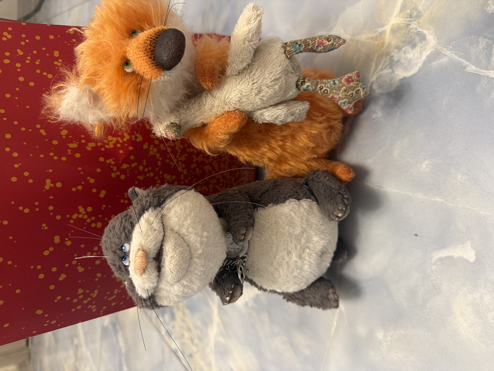 A plush cat and a plush fox are sitting next to each other. The cat is grey and white and has a surprised expression with its mouth open. It is wearing a metal charm around its neck. The fox is orange and white and is holding a light-colored plush in its arms. The fox is wearing patterned leggings. They are sitting on a light blue surface that appears to be a countertop or table. Behind them is a red object with yellow polka dots.