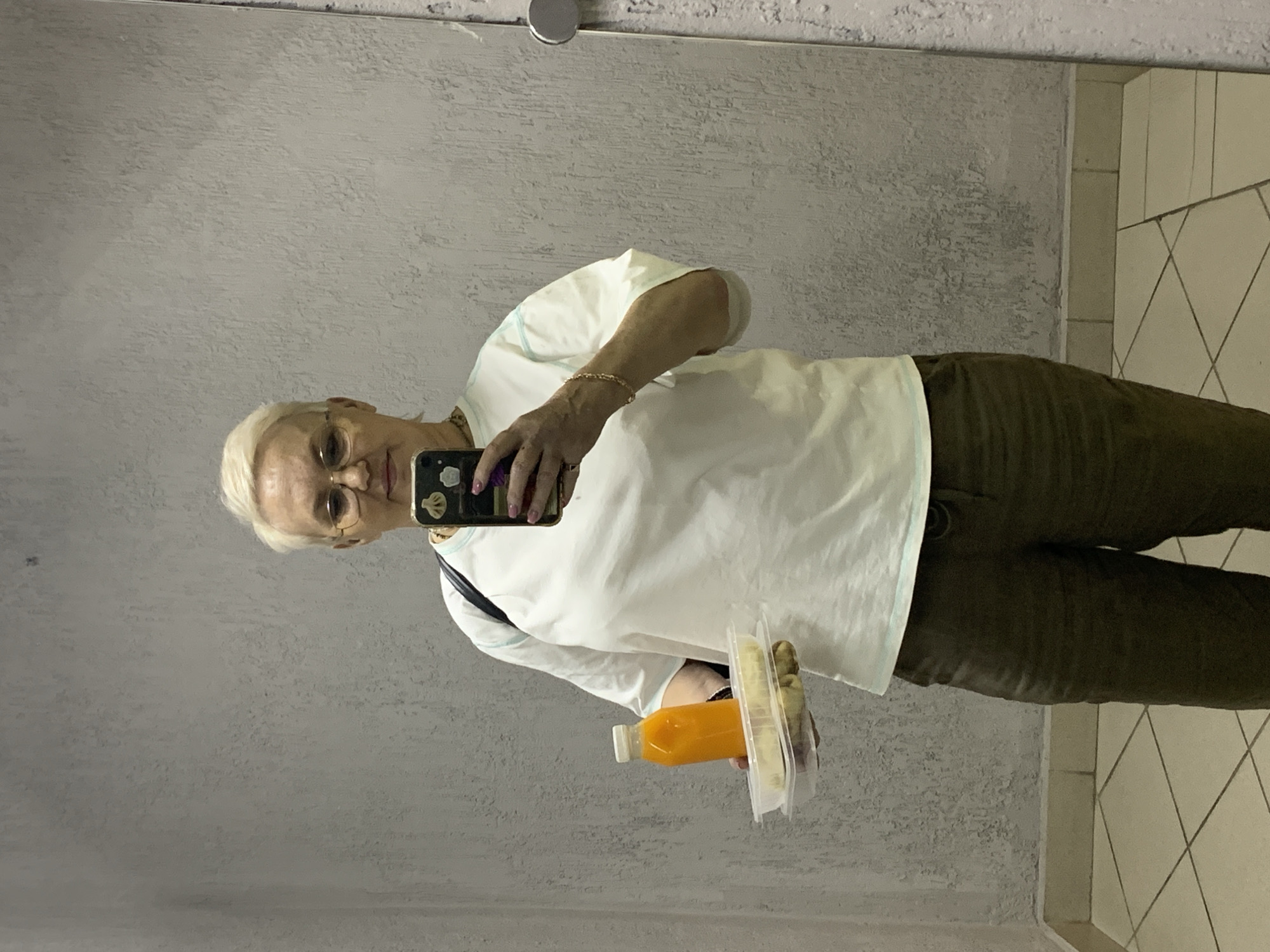 An older woman is taking a selfie in a bathroom mirror. She has short blonde hair and wears glasses, a white shirt, and olive green pants. She is holding a smartphone in one hand and a bottle of orange juice and a packaged food item in the other. The bathroom has a grey wall and white tiled flooring. The location is Chaikovsky, Russia.