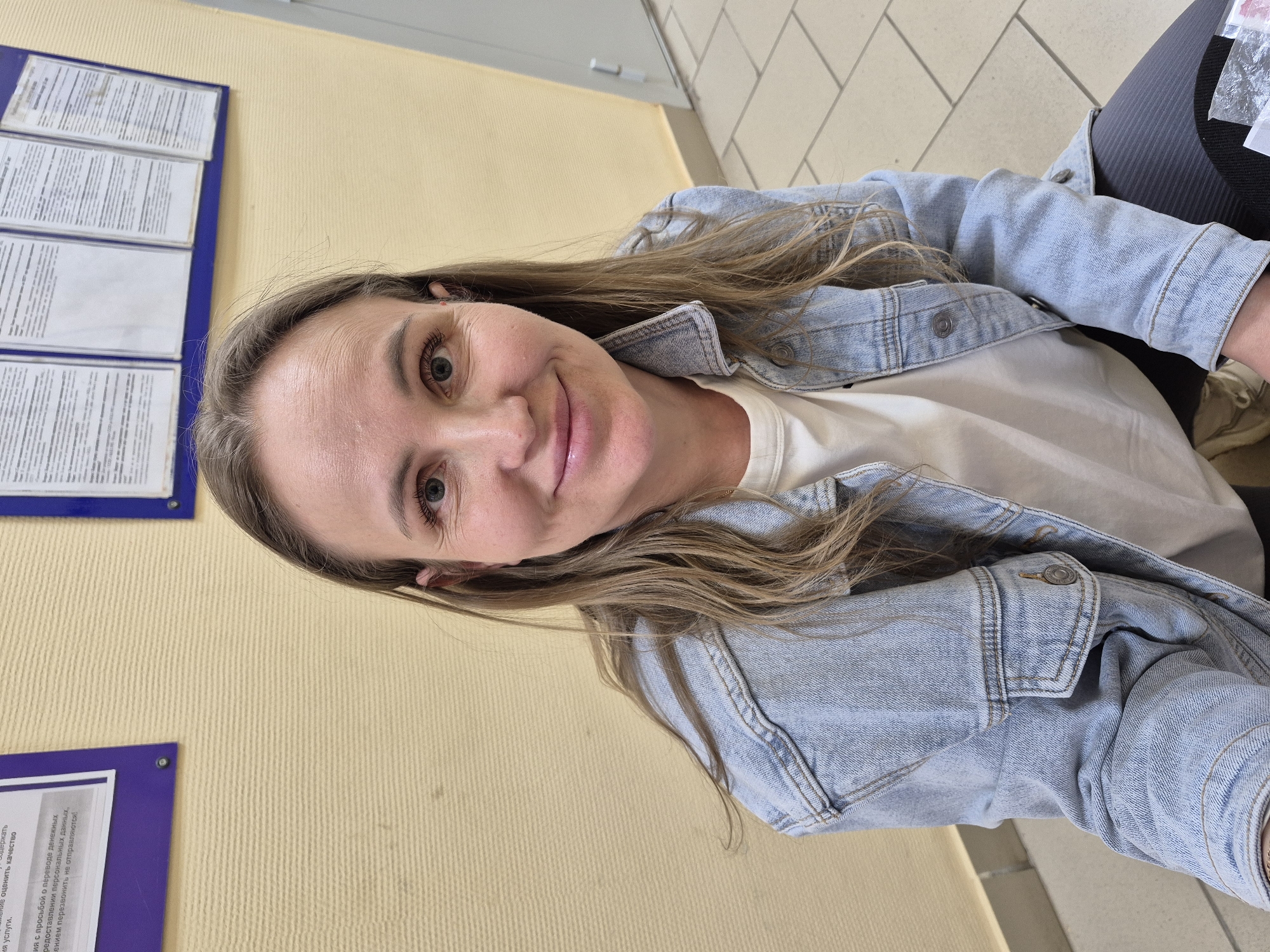 A woman is looking at the camera. She has fair skin, hazel eyes, and long brown hair. She's wearing a light blue denim jacket over a white shirt. She appears to be indoors, possibly in an office or waiting area. The background includes a beige wall with two informational posters framed in blue. The posters contain text in what appears to be Russian. The environment seems sterile and functional. The bottom left corner of the image shows a partially visible text that reads, "...с просьбой о переводе денежных...". The image appears to have been taken indoors in Chaikovsky, Russia.
