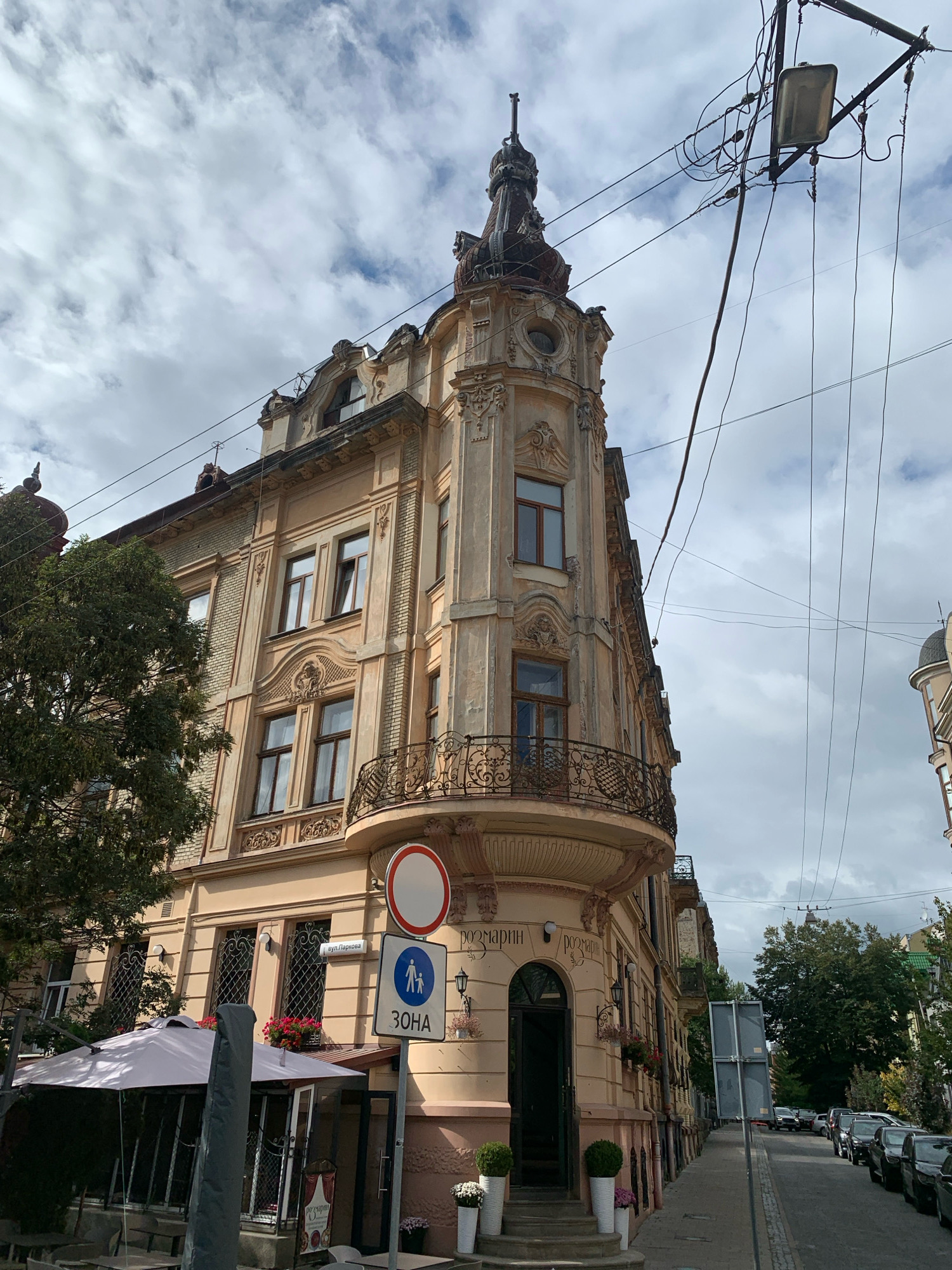 The image shows a corner building on a street in Lviv, Ukraine. The building is made of light-colored stone and has ornate architectural details, including balconies with wrought-iron railings and decorative carvings around the windows. The building's facade features a mixture of light yellow and beige tones. Above the building, a tangle of electrical wires and a street lamp are visible against a cloudy sky.
On the ground level, there is an entrance to the building with a dark-colored door, flanked by two white cylindrical planters containing greenery. Next to the entrance, a white umbrella shelters an outdoor seating area, suggesting a cafe or restaurant. A sign with Cyrillic text, "РОЗМАРИН," is visible above the entrance, likely indicating the name of the establishment.
Several street signs are present. One is a circular red-and-white traffic sign, and another is a blue sign with a white pedestrian icon and the Cyrillic word "ЗОНА" (ZONE), indicating a pedestrian zone. A street sign with Cyrillic text, "вул.Паркова," is also visible.
The street itself is paved with cobblestones, and a row of parked cars is lined up along the right side of the road. Trees and more buildings can be seen further down the street. The overall scene depicts a typical urban environment in Lviv, with a blend of historical architecture and modern life.