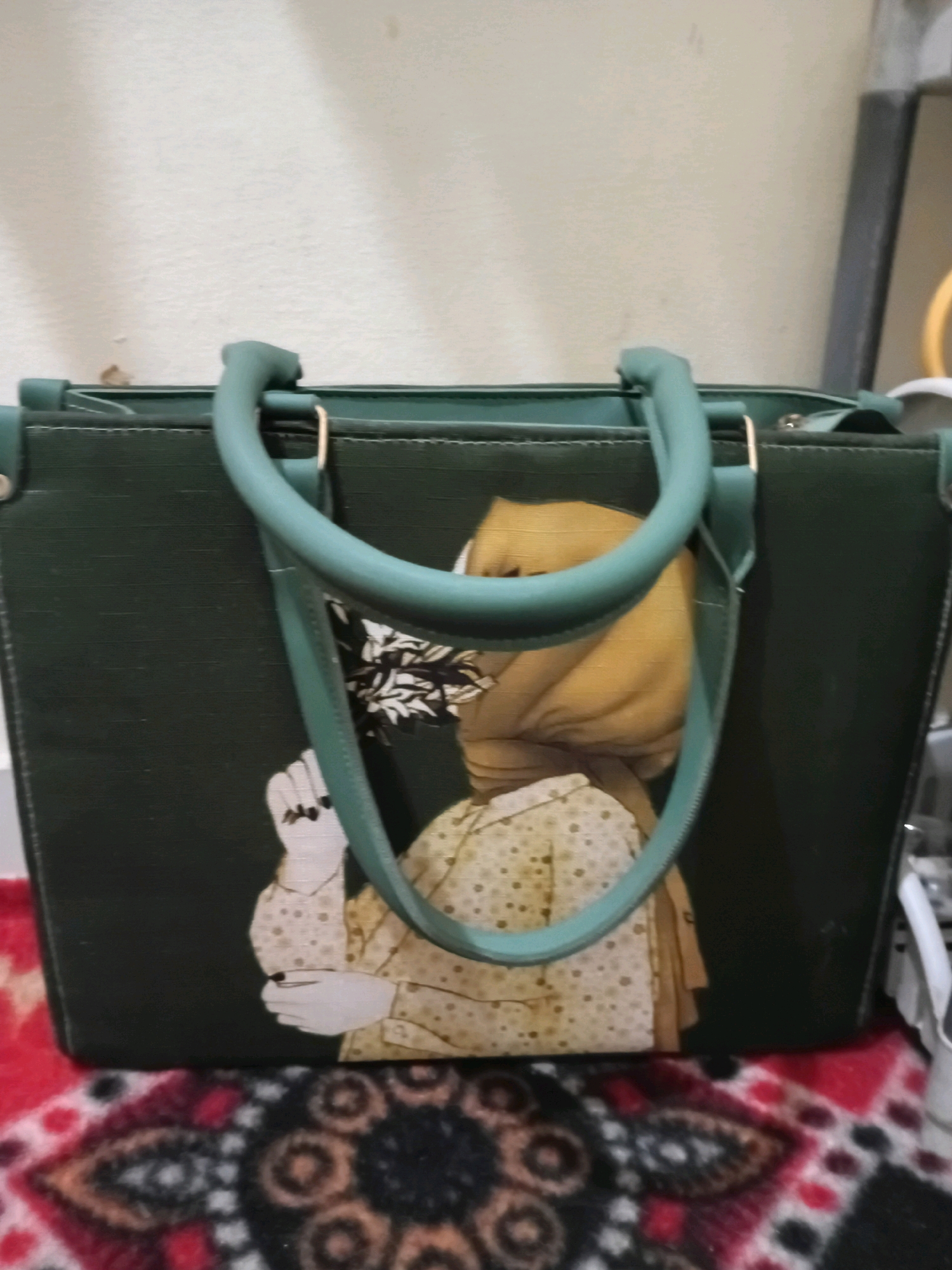 A dark green tote bag with teal handles is displayed against a light-colored wall. The bag features a printed image of a person wearing a yellow hijab and a patterned shirt. The person's left hand is visible, holding a bouquet of flowers. The bag rests on a patterned rug. The scene is indoors, with a portion of a framed object visible in the background. There is no discernible location in Muzaffargarh, Pakistan.