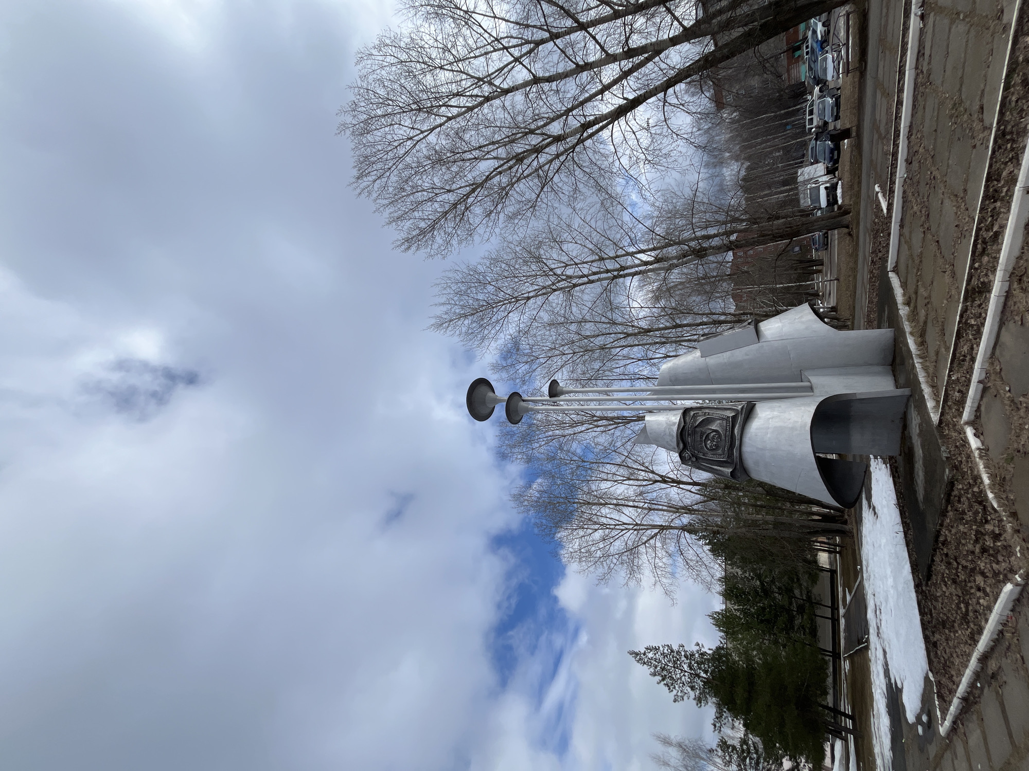 The image shows a monument located in the city of Chaikovsky, Russia, under a partly cloudy sky. The monument, made of a silver-colored metal, features a structure with three horn-like projections extending upwards. The base of the monument has a decorative relief. Bare trees surround the monument. In the background, there is a parking lot with several parked vehicles. Some remaining snow is present on the ground near the monument. The photograph is taken from a low angle, looking upwards towards the sky.