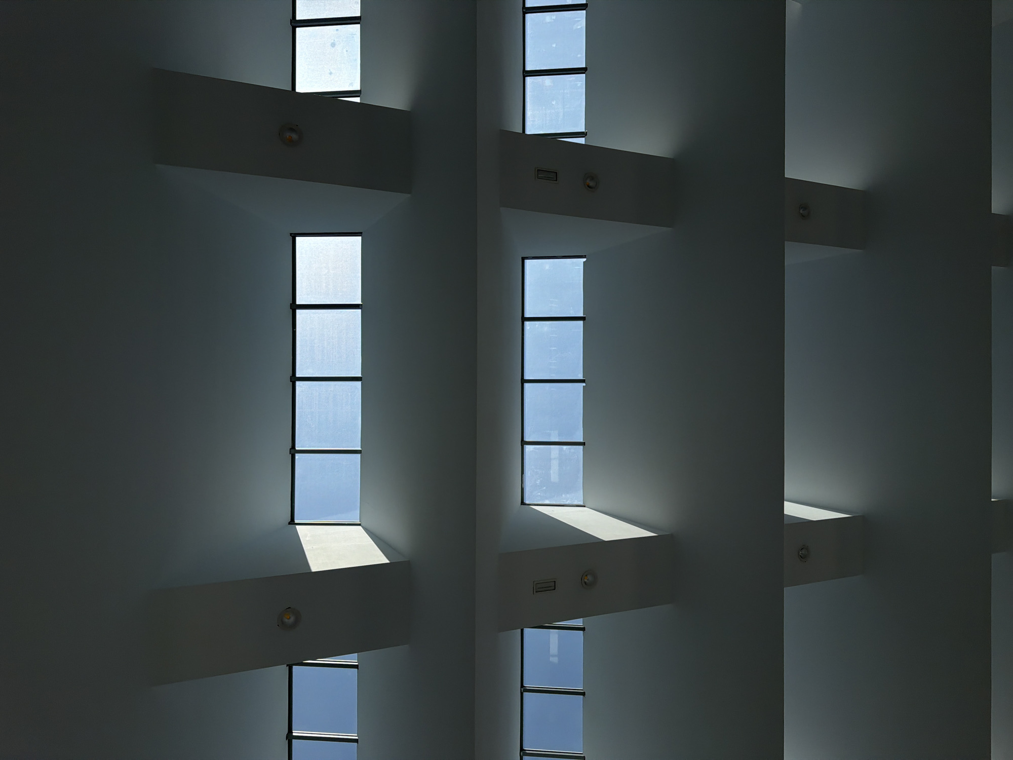 The image displays a ceiling with three horizontal rows of windows, characteristic of modern architecture. The windows are rectangular and framed by dark material, with the sky visible through them. They are arranged in pairs, separated by white beams that cast shadows on the ceiling. In the middle row, there are also circular lights and rectangular vents visible on the beams. The scene is likely indoors, possibly in a public building or office space, given the architectural features. There are no people or distinct objects other than the architectural elements. The lighting suggests it is daytime, with natural light streaming through the windows. The image is framed to emphasize the geometric patterns of the ceiling, beams, and windows.