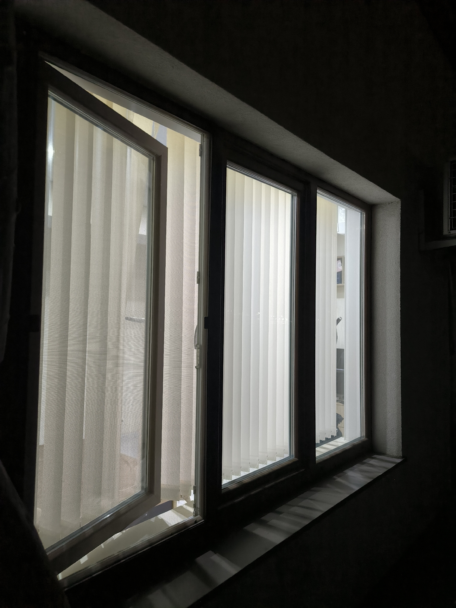 The image depicts an interior view of a window with vertical blinds. The window has multiple panes, with the leftmost pane slightly ajar, tilted inwards at the top. The blinds are a light, neutral color, creating a striped pattern across the visible window area. The window is set into a textured, light-colored windowsill, which is illuminated by light filtering through the blinds, casting striped shadows onto the dark wall below. The surrounding environment is dimly lit, suggesting an indoor setting at night or in low light conditions. To the far right, a portion of an air conditioning unit is visible mounted high on the wall. The scene is captured from a low angle, looking upwards towards the window. There are no people or animals present in the image.