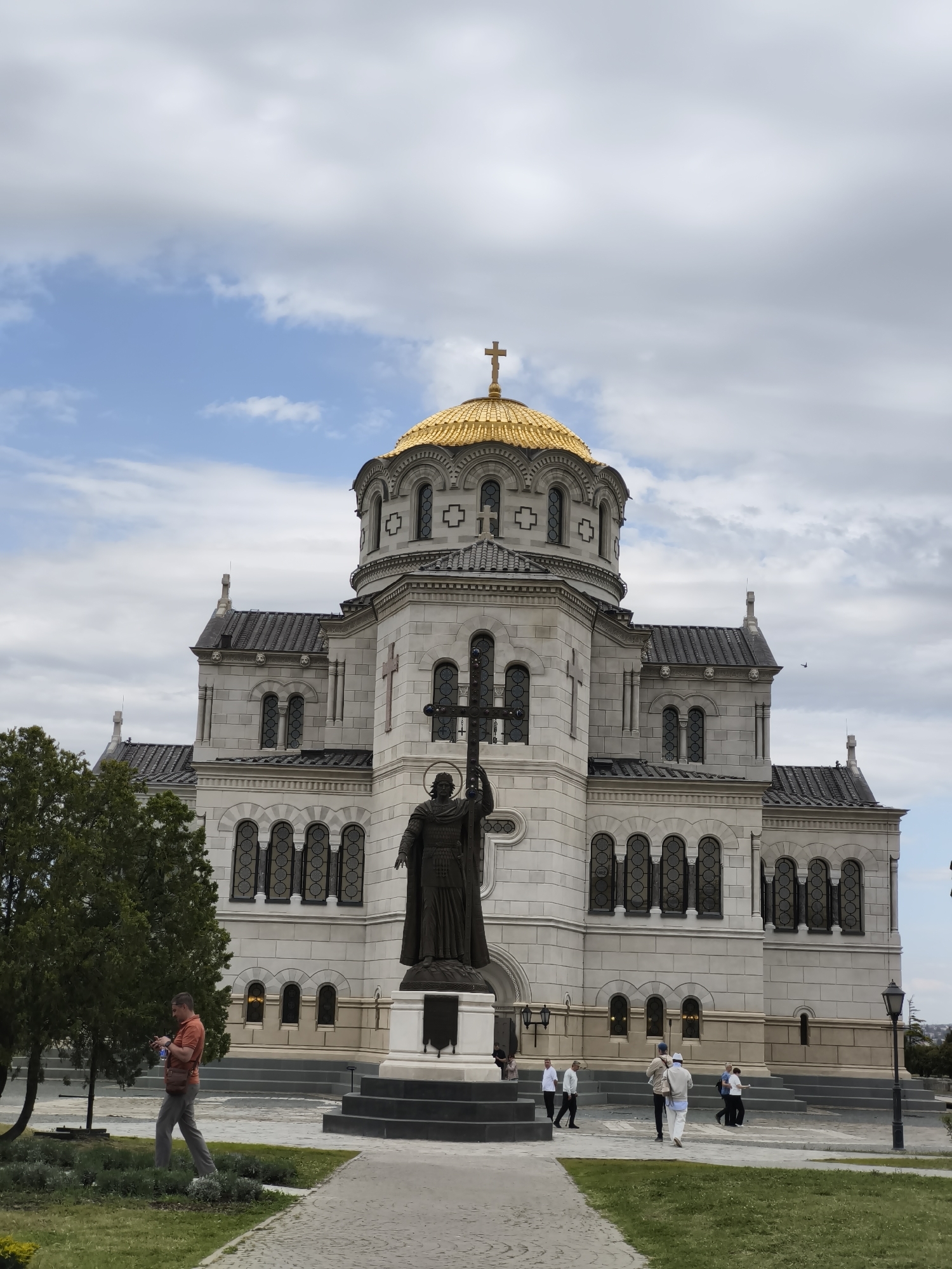 The image shows the St. Vladimir Cathedral in Sevastopol, Ukraine. The cathedral is made of white stone and has a large golden dome topped with a cross. In front of the cathedral is a bronze statue of a man holding a cross. People are walking around the square in front of the cathedral. A man in an orange shirt is walking on the lawn to the left of the cathedral. The sky is cloudy with patches of blue.
