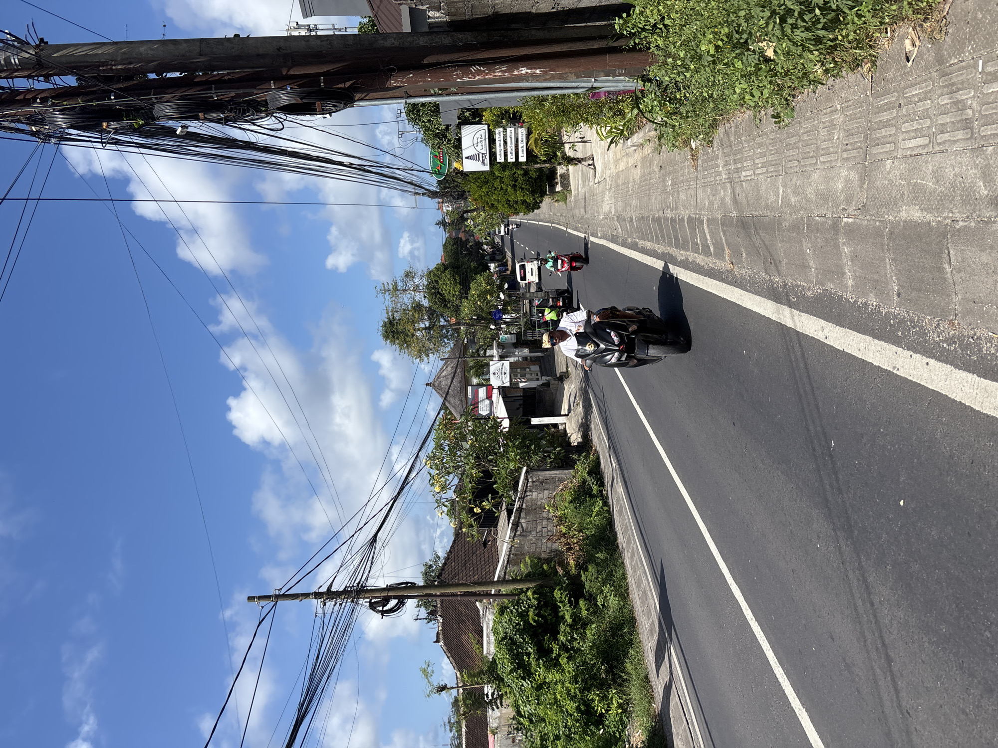 The image shows a street scene in Kuta Selatan, Indonesia. A paved road curves gently through the frame, marked by a white line. Motorbikes are the primary vehicles on the road, with one prominently in the foreground carrying two people. The rider wears a white shirt. Overhead, a network of power lines stretches between wooden utility poles on either side of the road. Buildings and lush green vegetation line the sides of the street. A sign displays business information. The sky is visible, displaying blue hues and scattered clouds. The photograph is oriented in a portrait perspective, causing the road to appear vertically aligned.