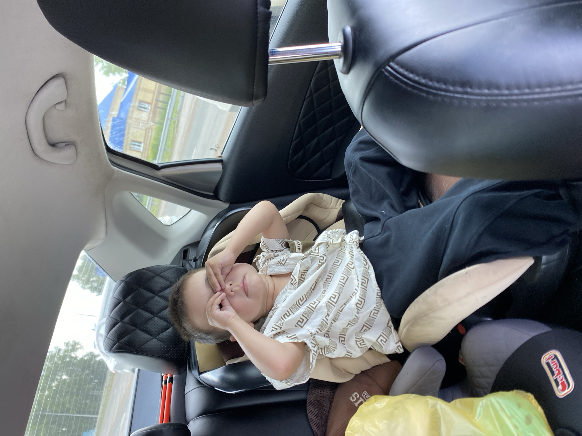 A young child is seated in a car seat in the back of a vehicle, looking tired with their eyes covered by their hands. The child is wearing a white patterned shirt and dark pants. In the background, a building with a blue roof is visible through the car window, alongside trees and a road. The interior of the car, including the seats and headrests, is visible. The scene appears to be within the city of Kushnarenkovo, Russia.