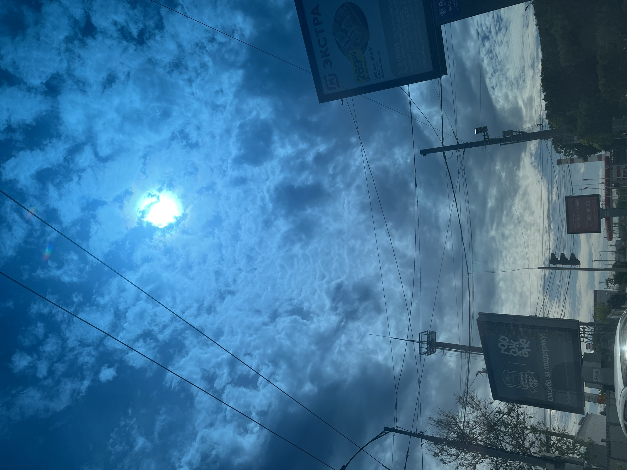 The image captures an upward view of a daytime urban street scene in Krasnodar, Russia, dominated by a dynamic sky and overhead infrastructure. The sun, partially obscured by scattered clouds, shines brightly in the upper left quadrant, creating a strong lens flare and a subtle rainbow effect. The sky transitions from a clear, vivid blue near the sun to a mix of white and darker, textured clouds across the rest of the frame, suggesting variable weather conditions.
Numerous black utility wires crisscross the expanse of the sky, connected to several visible utility poles. A pole on the right side supports a traffic light assembly, and further right, the dark green canopy of trees is visible against the sky. A bare tree branch extends from the lower right.
Two large billboards are prominent. The one in the upper right displays text in Cyrillic, reading "М ЭКСТРА" with a price "269⁹⁹", advertising "ШАШЛЫК СВИНОЙ В МАЙОНЕЗЕ ПОЛУФАБРИКАТ, 1 кг" available from "3-9 ИЮНЯ", along with a partial phone number "+7(861)12...". Another billboard in the lower right features the phrase "спасибо за поддержку!" and "МЕСТО СВОБОДНО" with a contact number "801 85 00". A small part of a white vehicle, likely a car, is visible in the bottom right corner, reflecting light. The overall scene depicts a typical city street environment, bustling with infrastructure but devoid of direct human presence or specific activity.