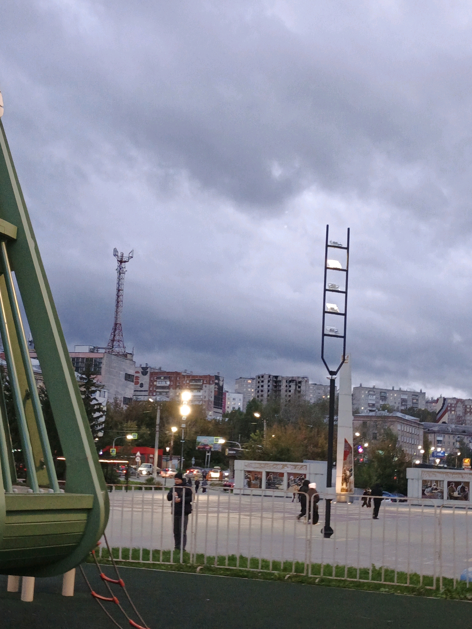 The image depicts a wide, overcast view of a city square in Zlatoust, Russia. In the foreground, a portion of a green playground structure is visible on the left, with red ropes attached. A white metal fence runs horizontally across the middle ground, behind which several individuals are observed. One person in a dark jacket and pants stands near the fence on the right, holding what appears to be a dark object. Further back, other figures are walking or standing in the square. Beyond the square, a street with moving vehicles and streetlights is visible, leading towards a backdrop of multi-story buildings. A prominent red and white telecommunications tower stands tall in the mid-background, to the left of a vertical art installation resembling a ladder with illuminated elements. The sky is filled with heavy, grey clouds.