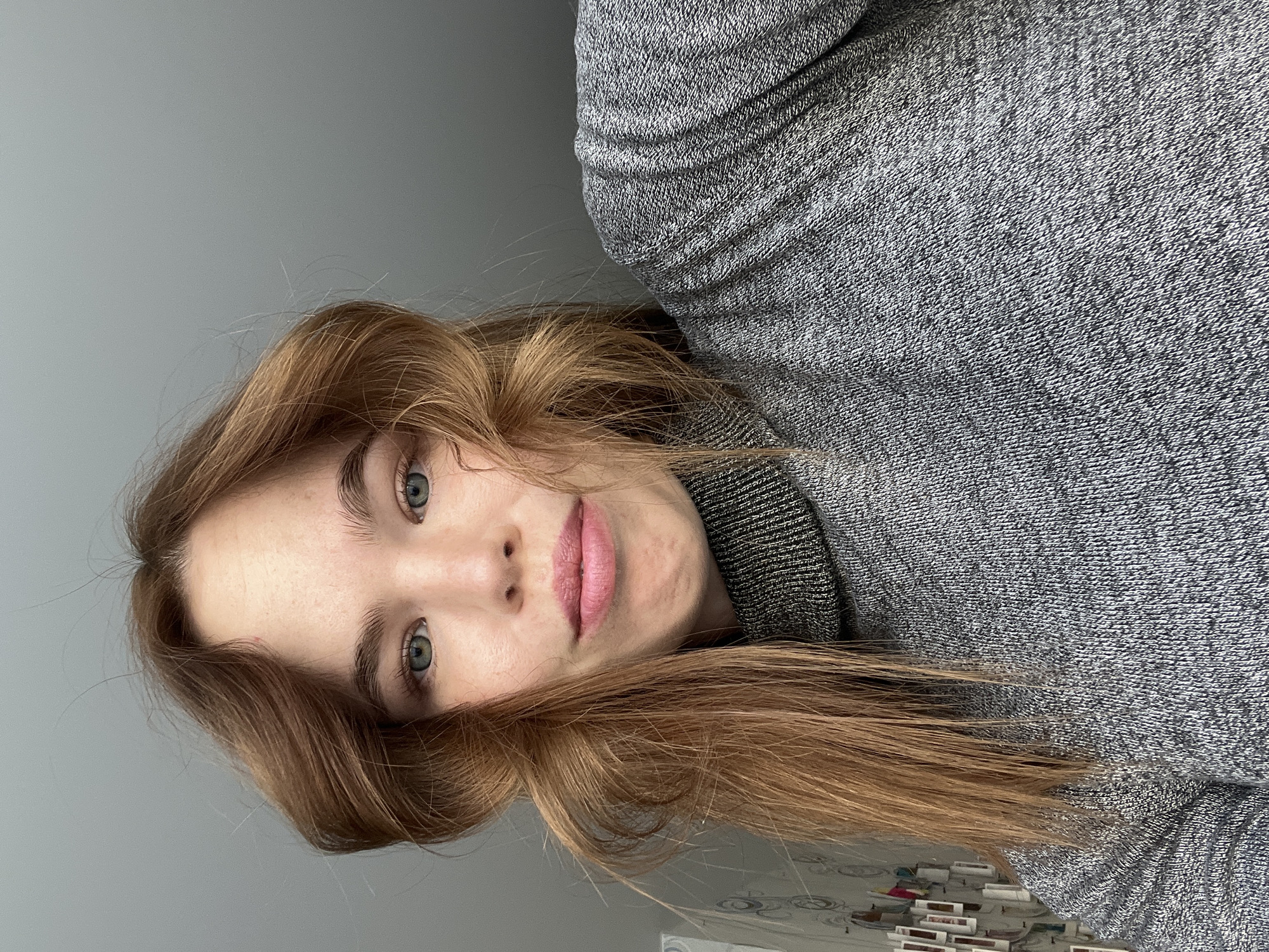 A photograph of a person with light brown hair and light-colored eyes, wearing a grey knitted turtleneck sweater. The person is positioned close to the camera, with the face slightly tilted. The background is a plain grey wall. There are indistinct items visible at the bottom edge of the photo, resembling small objects or books. There is no indication of Chaikovsky, Russia or specific location.