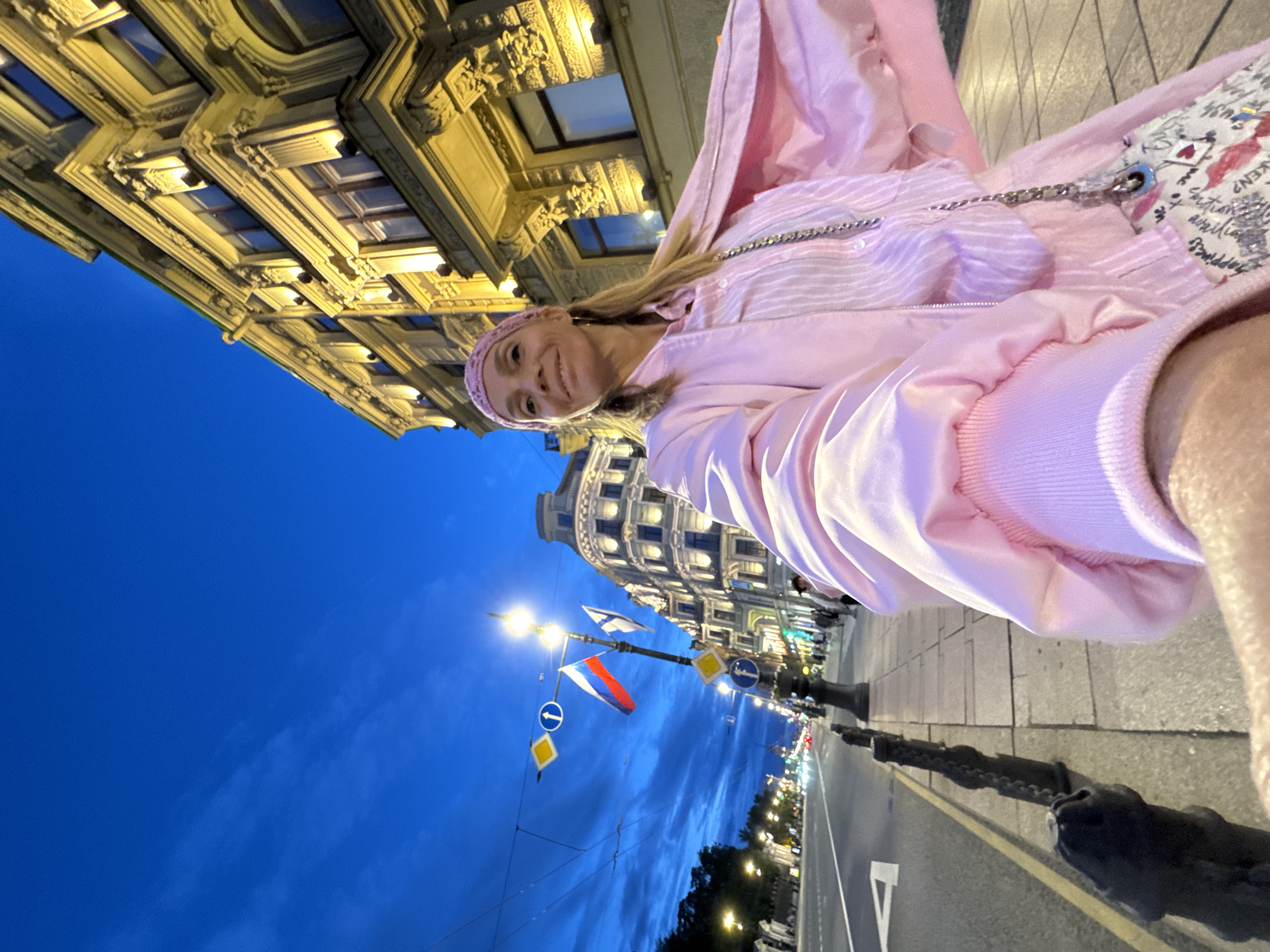 A woman, wearing a pink bandana, a pink collared shirt, and a pink bomber jacket, takes a selfie on a city street at dusk. She is standing on a sidewalk next to a street with cars and buildings on the right. The sky is a deep blue with clouds. Two flags, one Russian and one Finnish, fly from a lamppost. The buildings are illuminated by yellow lights.