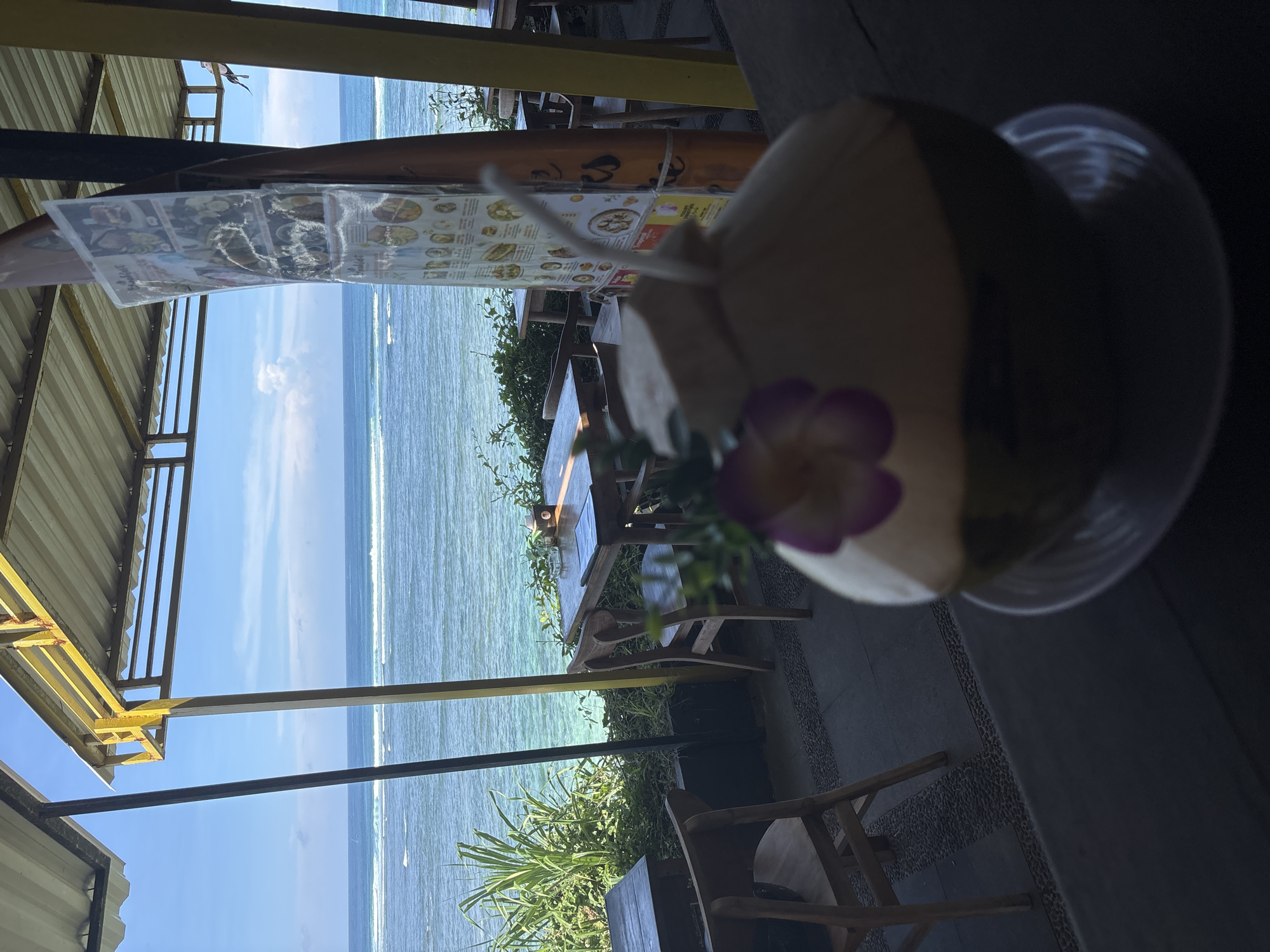 The image depicts a view from a seaside establishment in Kuta Selatan, Indonesia. The ocean is visible, and waves are breaking near the shore. The scene is partially framed by the structure of the open-air venue, which includes a corrugated metal roof and structural supports. A menu is affixed to the structure. In the foreground, there's a coconut drink served in a plastic container, adorned with a purple flower. The drink is placed on a table, and chairs are nearby, suggesting a seating arrangement for customers. The environment includes tropical vegetation.
