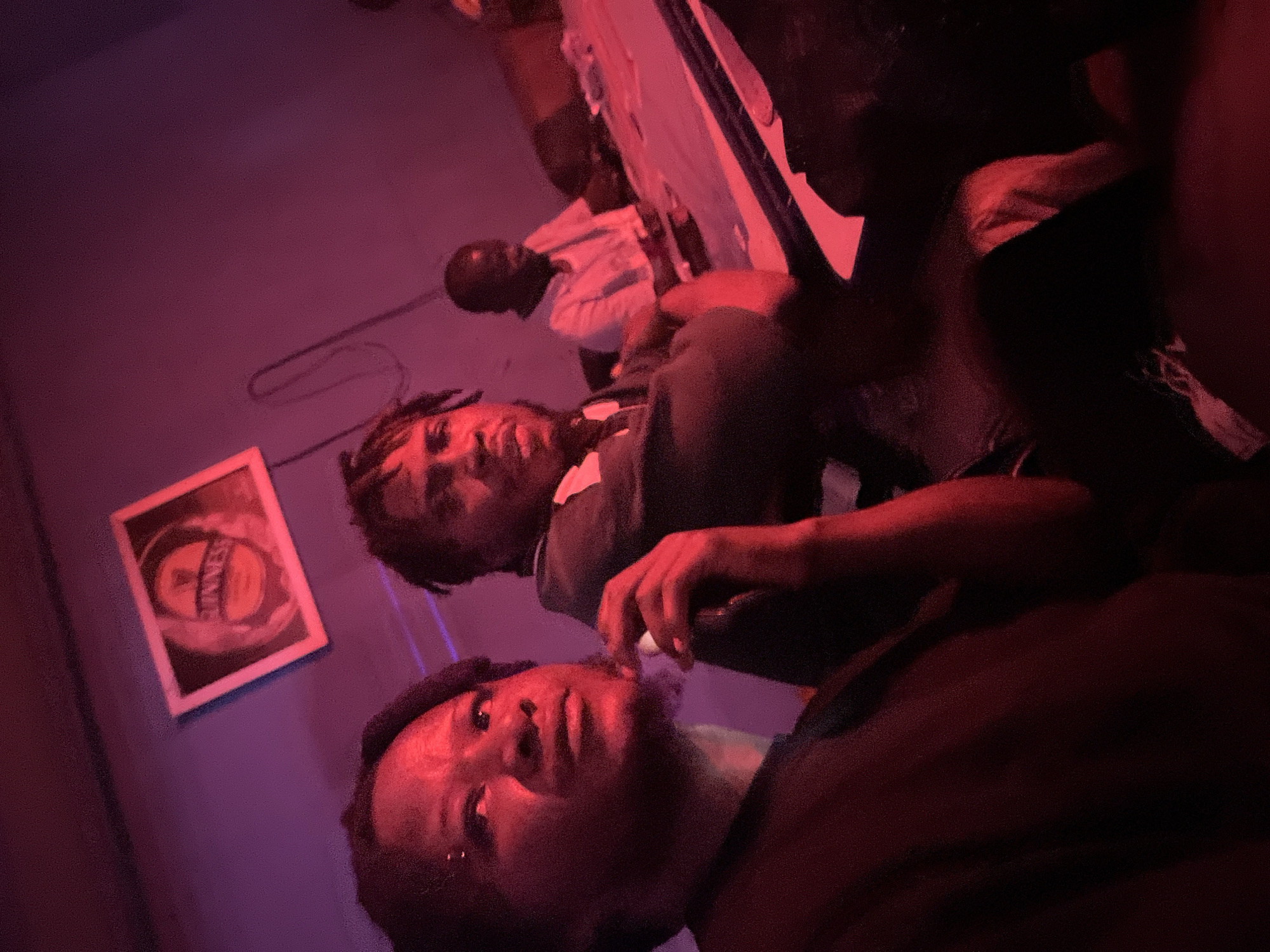 In a dimly lit room, possibly a bar or lounge in Jos, Nigeria, three individuals are present. The image is illuminated by a strong red light, casting a reddish hue over the scene and obscuring some details. In the foreground, a person with dark skin and dreadlocks is partially visible, their face turned to the left, looking upwards. They are wearing a dark t-shirt. To their right and slightly behind, another person with dark skin and dreadlocks is more clearly visible, facing forward and looking towards the right. This individual is wearing a dark green jacket with a white collar. Further back, a third person with dark skin is seated, wearing a white t-shirt and looking down at a table. The background features a wall with a framed picture of a Guinness beer logo. A cable hangs from the wall above and behind the person in the foreground. The individuals appear to be seated at a table, though the table's surface is mostly obscured by shadows and the red lighting. The overall atmosphere is informal and social.