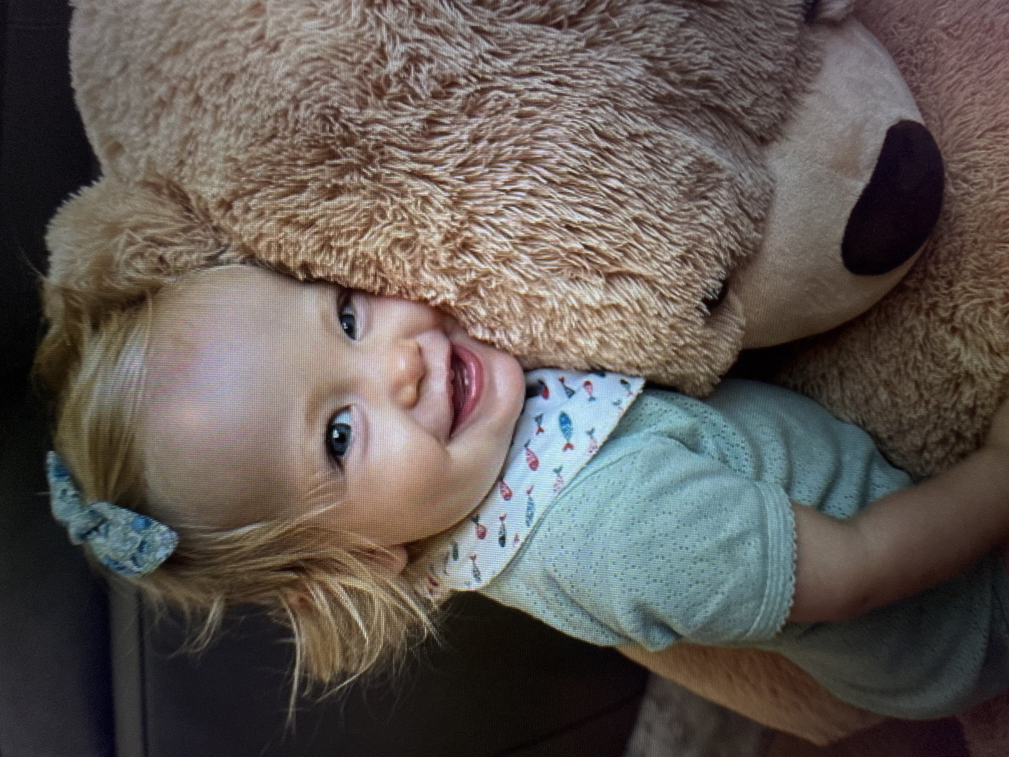 A joyful baby or toddler with light blonde hair and bright blue eyes is depicted in a close-up, chest-up portrait, playfully nestled against a large, fluffy brown teddy bear. The child is smiling widely with an open mouth, showing their tongue, and looking directly at the camera. Their head is positioned beneath the bear's head, which is visible on the right side of the frame with a distinct dark brown nose.
The baby wears a light blue or mint green long-sleeved top featuring a textured knit pattern. A white Peter Pan collar, adorned with a repeating pattern of small, colorful fish (including red and blue), adds a charming detail to the outfit. A small, light blue patterned bow is clipped into the child's hair on the left side of their head. The large teddy bear is made of soft, shaggy brown material, creating a cozy and comforting presence.
The background is dark and out of focus, indicating an indoor setting, likely a home environment. The lighting is soft and even, illuminating the subjects. The overall mood of the image is warm, affectionate, and cheerful, capturing a moment of happiness and comfort. There is no visible text in the image.