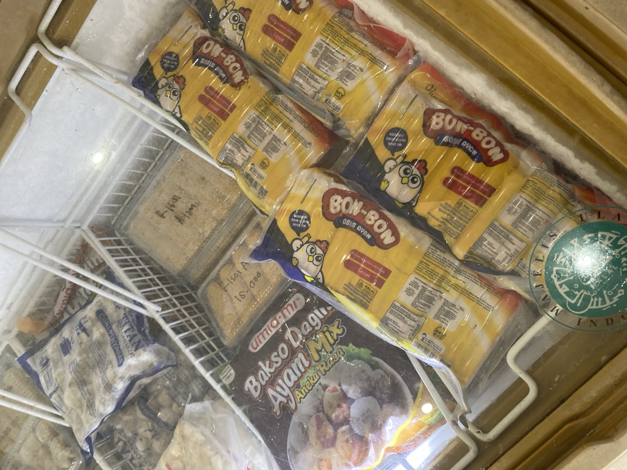 The image shows a close-up view of a freezer display case filled with frozen food items. The products are primarily packaged sausages and what appear to be frozen meat products. In the foreground, several packages of yellow sausage with cartoon chicken designs are visible, labeled "BON-BON" and "Sosis Ayam". To the left, a larger package of "Bakso Daging Ayam Mix Aneka Rasa" is displayed. In the background, there are other frozen food packages, some with visible text like "Risoles Ayam". A prominent green circular seal with Indonesian text, likely a halal certification, is partially visible in the lower right corner. The freezer case itself appears to be made of metal and glass, with white wire racks holding the products. The overall lighting suggests an indoor commercial setting, such as a supermarket or convenience store. The location is identified as Payatieng, Indonesia.