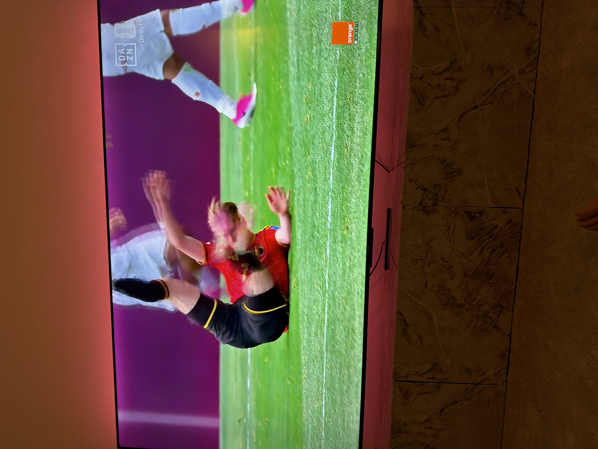 In the city of Barcelona, Spain, a television screen displays a soccer match. A female player in a red and yellow uniform is in mid-air, falling on a green grass field. Another player in a white uniform with pink shoes is visible in the background. The screen shows logos for DAZN and FIFA. The image is taken from a low angle, with the TV placed on a stand that is lit with pink lighting. The background is dark, suggesting the room is dimly lit.
