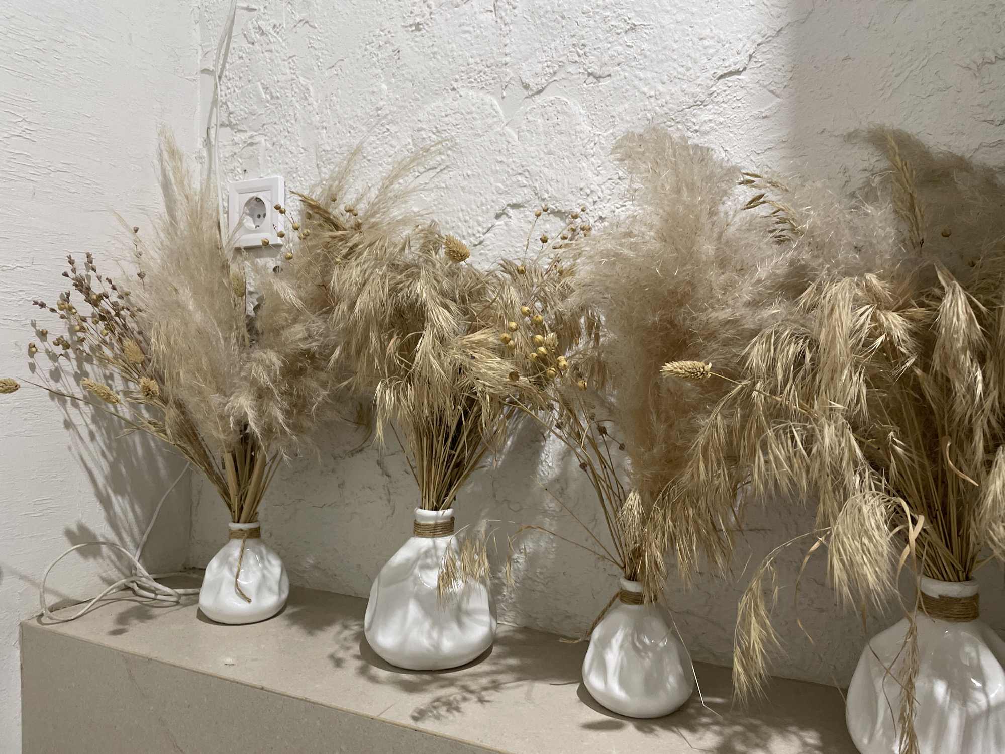 The image depicts a close-up, eye-level, indoor shot of several vases filled with dried ornamental grasses. The vases are white, with an irregular, bulbous shape, and each is loosely tied around the neck with twine. The dried grasses are light brown and beige, with feathery plumes and some seed heads. They are arranged in a way that creates a soft, airy texture.
The vases are placed on a flat, beige surface, possibly a shelf or countertop, which extends across the bottom of the frame. Behind the vases, a textured white wall is visible. On the upper left side of the wall, a white electrical outlet with two circular sockets is mounted. A white electrical cord is seen on the shelf, trailing from the direction of the outlet and loosely looping near the leftmost vase. The lighting appears to be natural, casting subtle shadows from the grasses onto the wall.
The scene is devoid of people and animals. The overall impression is one of rustic or bohemian decor, likely in a domestic or commercial interior setting. There is no identifiable information within the image to suggest it is located in the city of Khmelnytskyi, Ukraine.