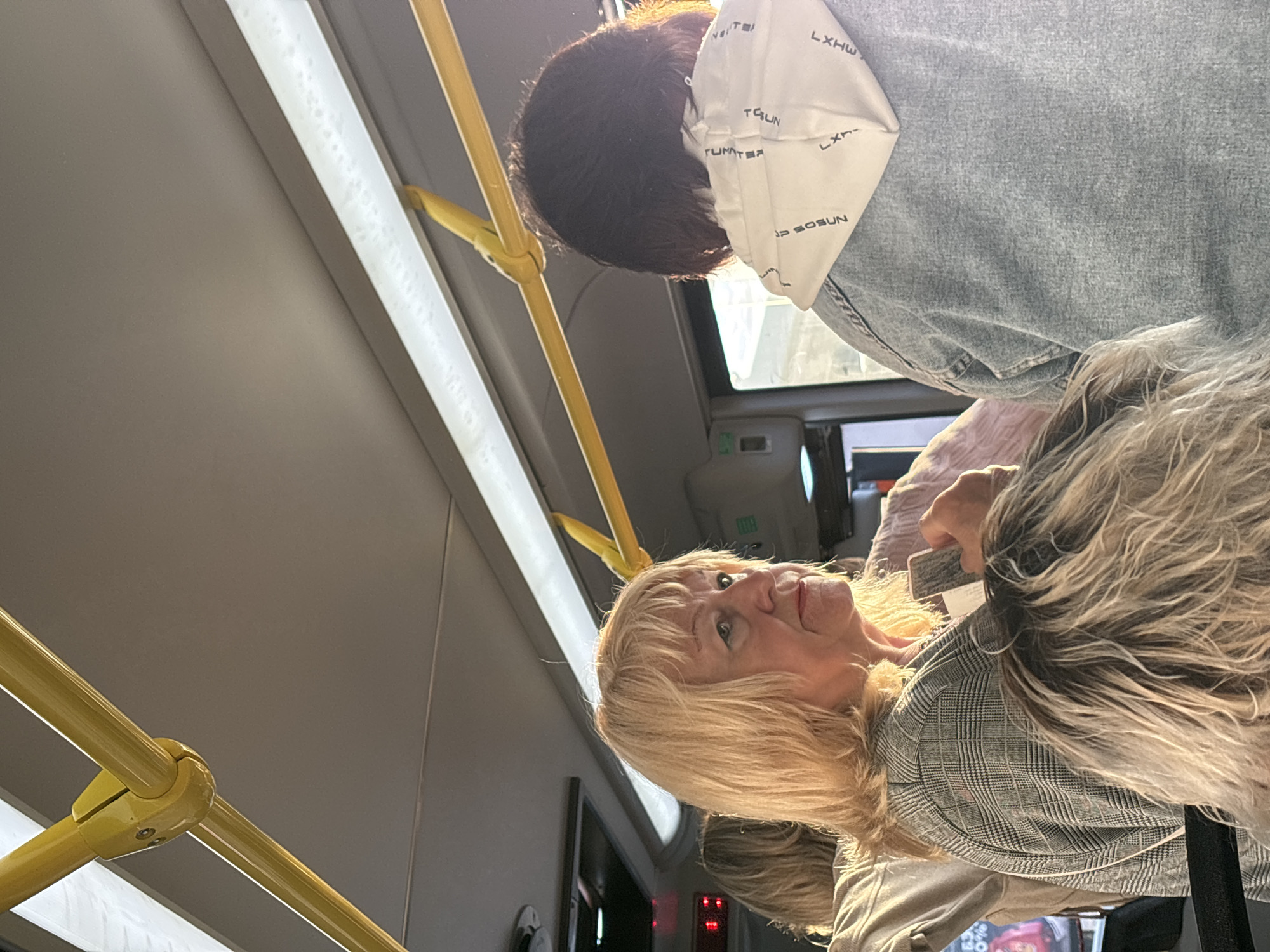 In Nizhny Novgorod, Russia, on a public transport vehicle, likely a bus or trolleybus, several passengers are visible. In the foreground, a woman with blonde hair and a gray checkered jacket is looking to her left, holding a rectangular object, possibly a ticket or phone. Her expression is neutral. Behind her, to the right, a person with dark hair wears a light-colored hooded sweatshirt with text printed on it, and a denim jacket. The interior of the vehicle is characterized by yellow handrails and fluorescent lighting. The scene is captured from a low angle, looking up towards the ceiling and handrails.