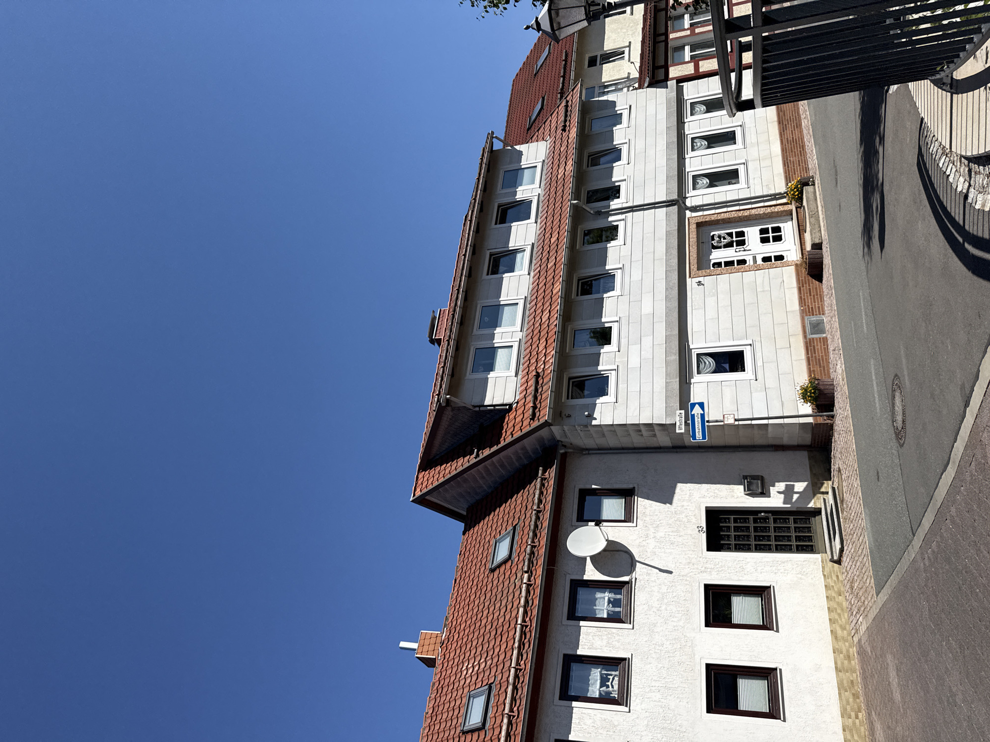 The image shows a residential street in Bad Sachsa, Germany, on a clear, sunny day. The scene features a row of traditional European houses with red tile roofs and white stucco or stone facades. Several windows, some with white blinds, are visible on each floor. A satellite dish is mounted on the exterior wall of one house, and a chimney extends from another roof. The street in the foreground is paved with asphalt, with a section of cobblestone or brickwork bordering the sidewalk. A street sign reads "Uffestraße" and indicates a one-way street with a blue arrow. A black metal railing and a street lamp are also present on the right side of the image. The overall impression is one of a quiet, well-maintained residential area.