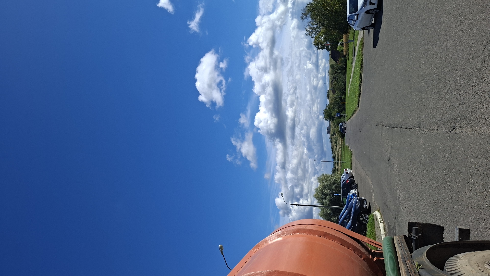 The image depicts a road scene under a bright blue sky with scattered white clouds. In the foreground on the left, a large orange cylindrical tank, likely part of a vehicle, is partially visible. A green hose extends from this tank. To the left of the road, several cars are parked in a lot. The road itself is paved and shows signs of wear, with cracks and potholes. Trees and green grass line the sides of the road, and in the distance, a few buildings and rolling hills are visible. The overall environment suggests an outdoor, possibly rural or suburban, setting. There are no people clearly visible in the image, and no actions are being performed. The location is identified as the city of Myadzyel, Belarus.