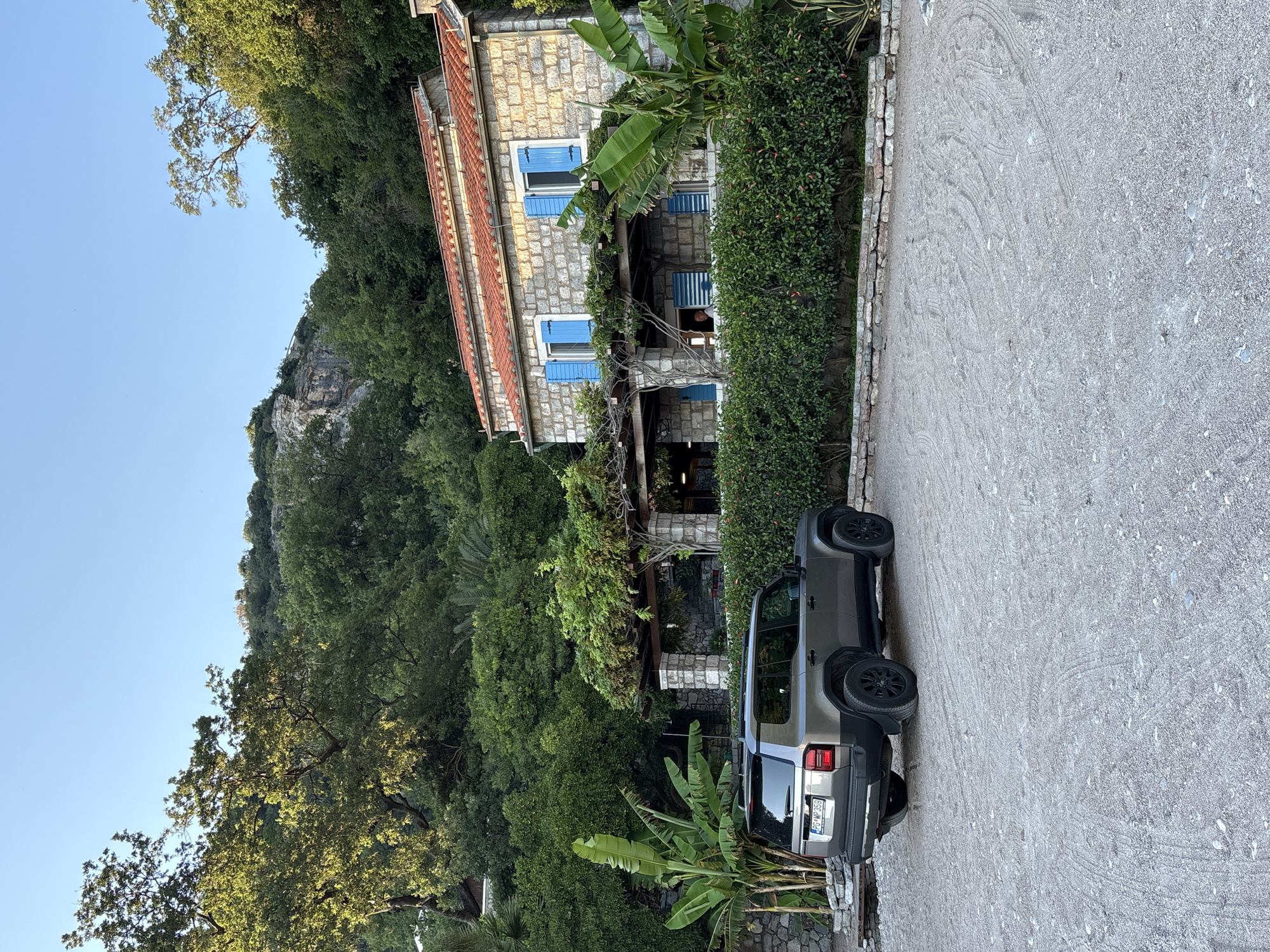 The image depicts a stone house with a red tile roof and bright blue shutters, situated amidst lush green foliage. A grey SUV is parked in a gravel lot in front of the house. The house features a covered patio area supported by stone pillars, with ivy and other plants climbing over the structure. Banana trees are visible to the left of the house, and a dense hedge lines the front of the property. The background consists of a steep, rocky hill covered in dense trees, characteristic of the natural landscape of Mirište, Montenegro. The sky is clear and blue, suggesting fair weather.
