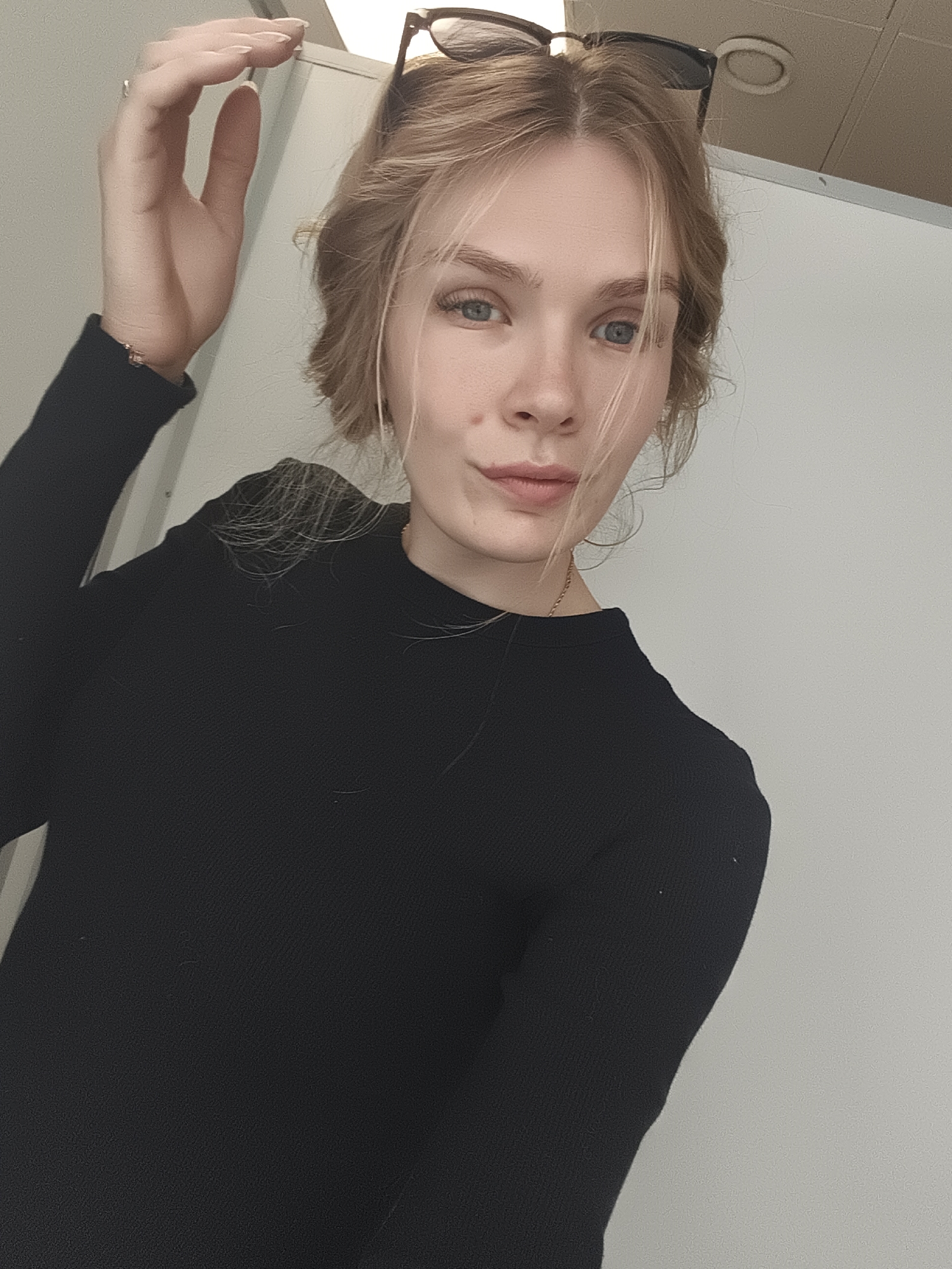 A young woman with fair skin and blonde hair poses indoors. She wears a black, long-sleeved top and has sunglasses perched on her head. Her right hand is raised near her head. The background consists of plain, light-colored walls. It is unknown if this image was taken in Izhevsk, Russia.