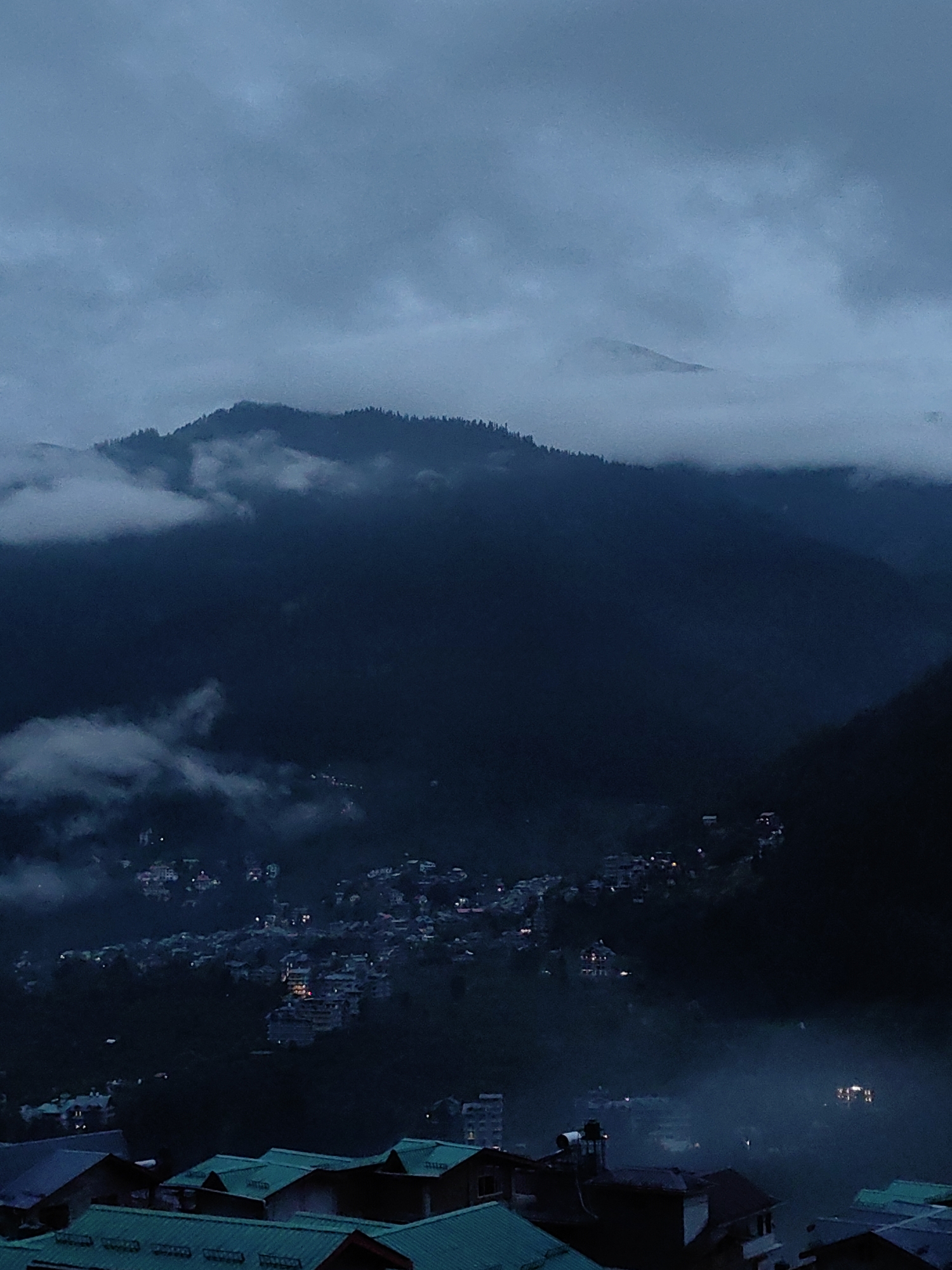 This is a vertical, medium-shot photograph of a mountainous landscape at dusk, featuring a small town nestled in a valley. The sky is overcast with dark, heavy clouds, giving the scene a somber and slightly ethereal mood. In the background, a large, dark mountain range dominates the upper portion of the frame, its peak partially obscured by mist and clouds. The slopes of the mountain are covered in dense evergreen trees.
In the middle ground, the town of Vashisht, India, is visible. The buildings are clustered together on the hillside, with numerous lights twinkling from windows and streetlights. The town appears to be built on a steep incline, with structures appearing at various levels. The lights suggest a population that is active even as darkness falls. Wisps of fog or mist are drifting through the town and across the slopes, softening the edges of the buildings and adding to the atmospheric quality of the image.
In the foreground, several rooftops of houses are visible. These are modern-looking structures with brightly colored, likely green, metal roofs. The roofs are angled and appear to be part of residential buildings. The perspective suggests the viewer is looking down from a higher vantage point, observing the town below. The overall impression is of a remote, mountainous community preparing for the night under a dramatic, cloudy sky.